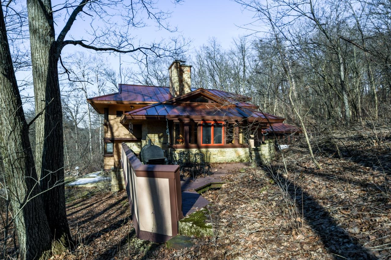 Stone-and-wood house with a copper roof, chimney, and raised deck among leafless trees.