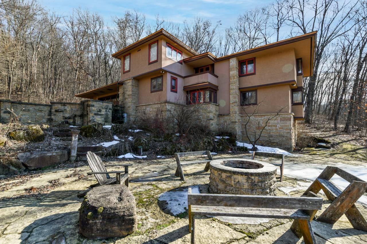 Multi-level home above a stone patio with a circular fire pit and wooden chairs.