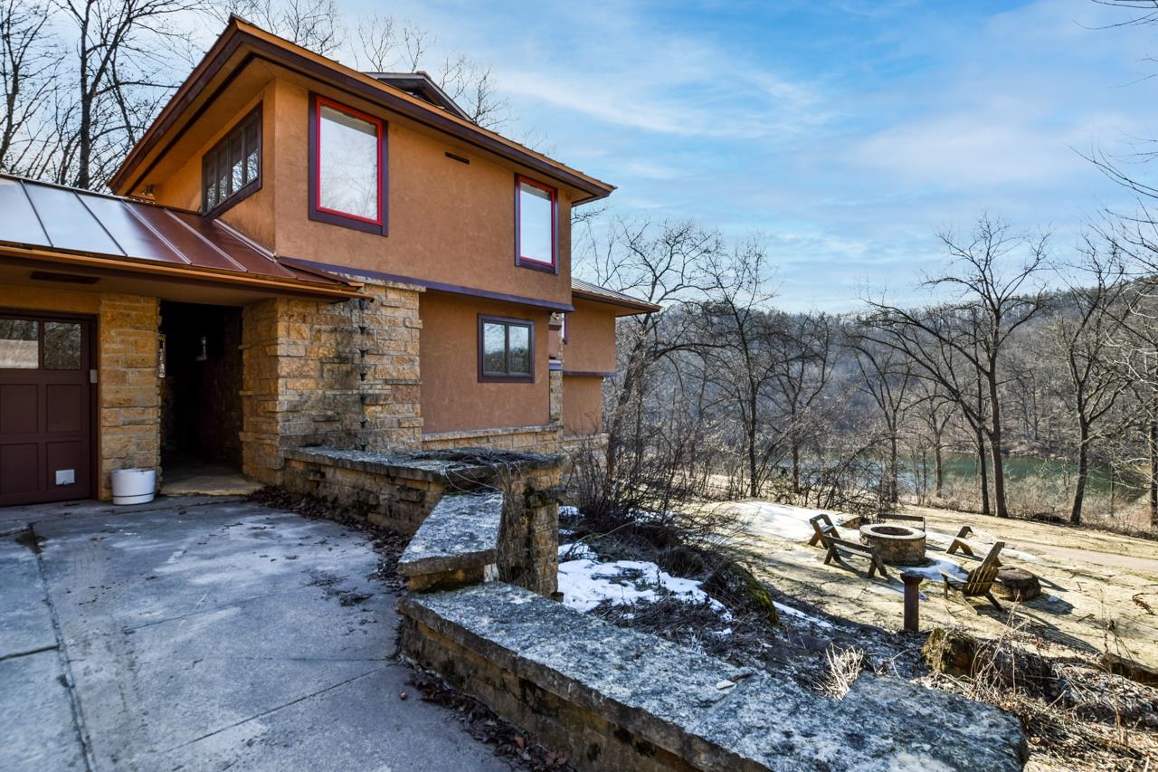 Brown two-story home with stone accents, a fire pit seating area, and water visible through leafless trees.