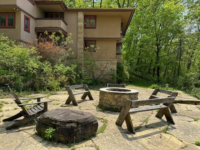 Multi-level home beside a wooded patio with a circular stone fire pit and wooden benches.
