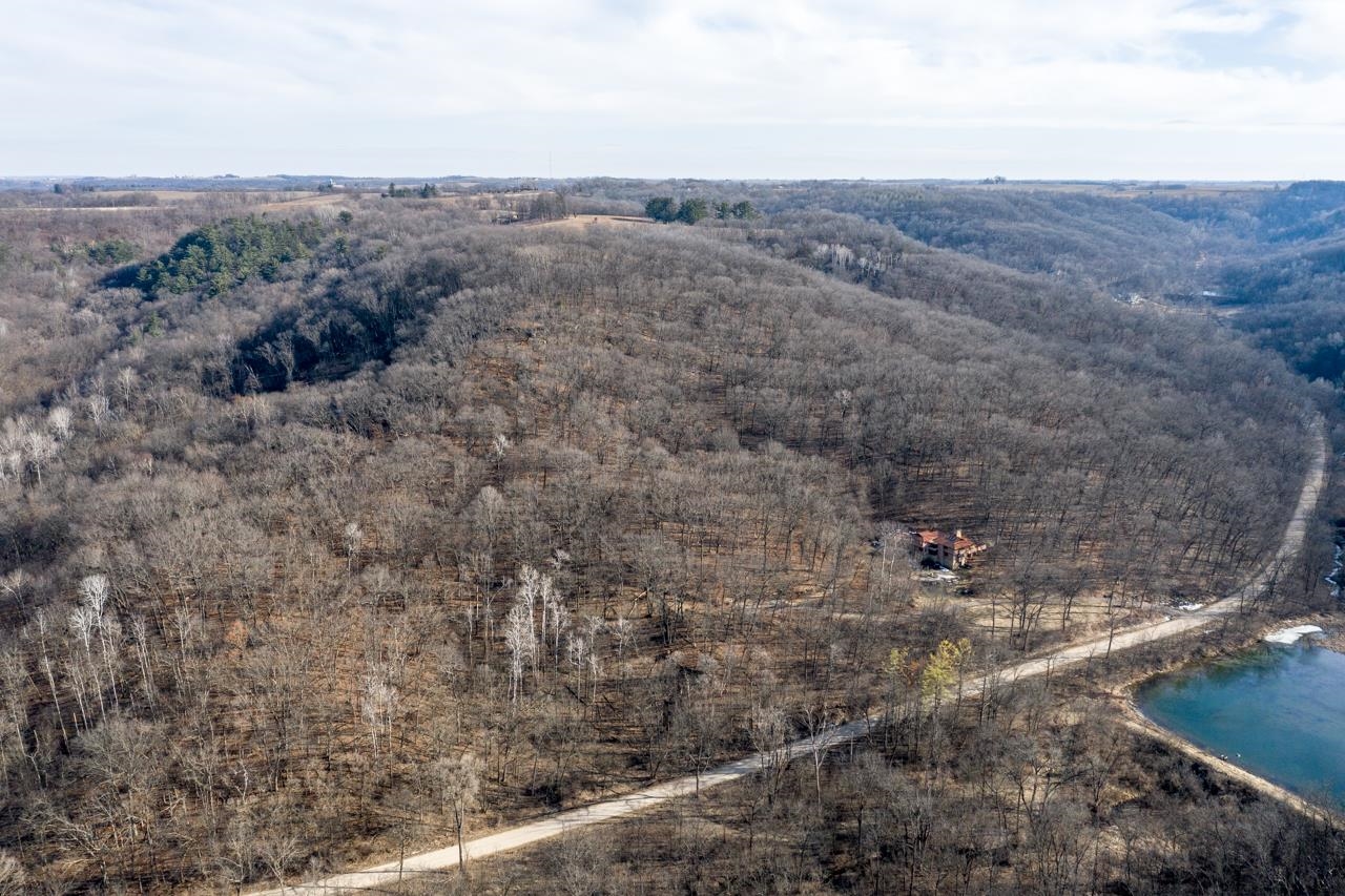 Aerial view of wooded hills with a house, winding dirt road, and pond.