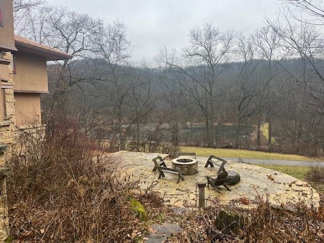 Stone patio with a fire pit and chairs overlooking leafless trees and a wooded hillside.
