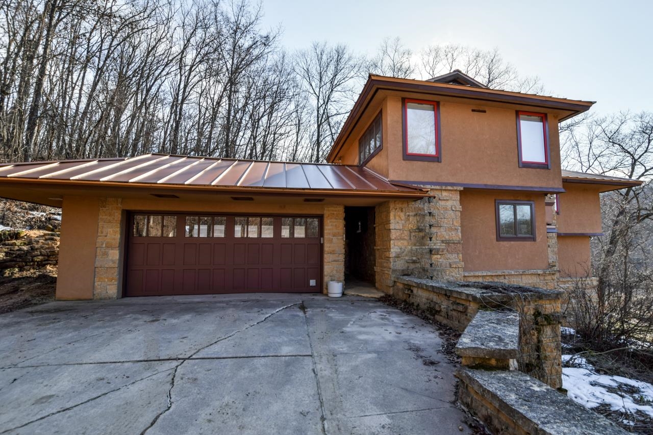Two-story tan home with stone accents, copper-colored roof, and a wide garage door amid leafless trees.