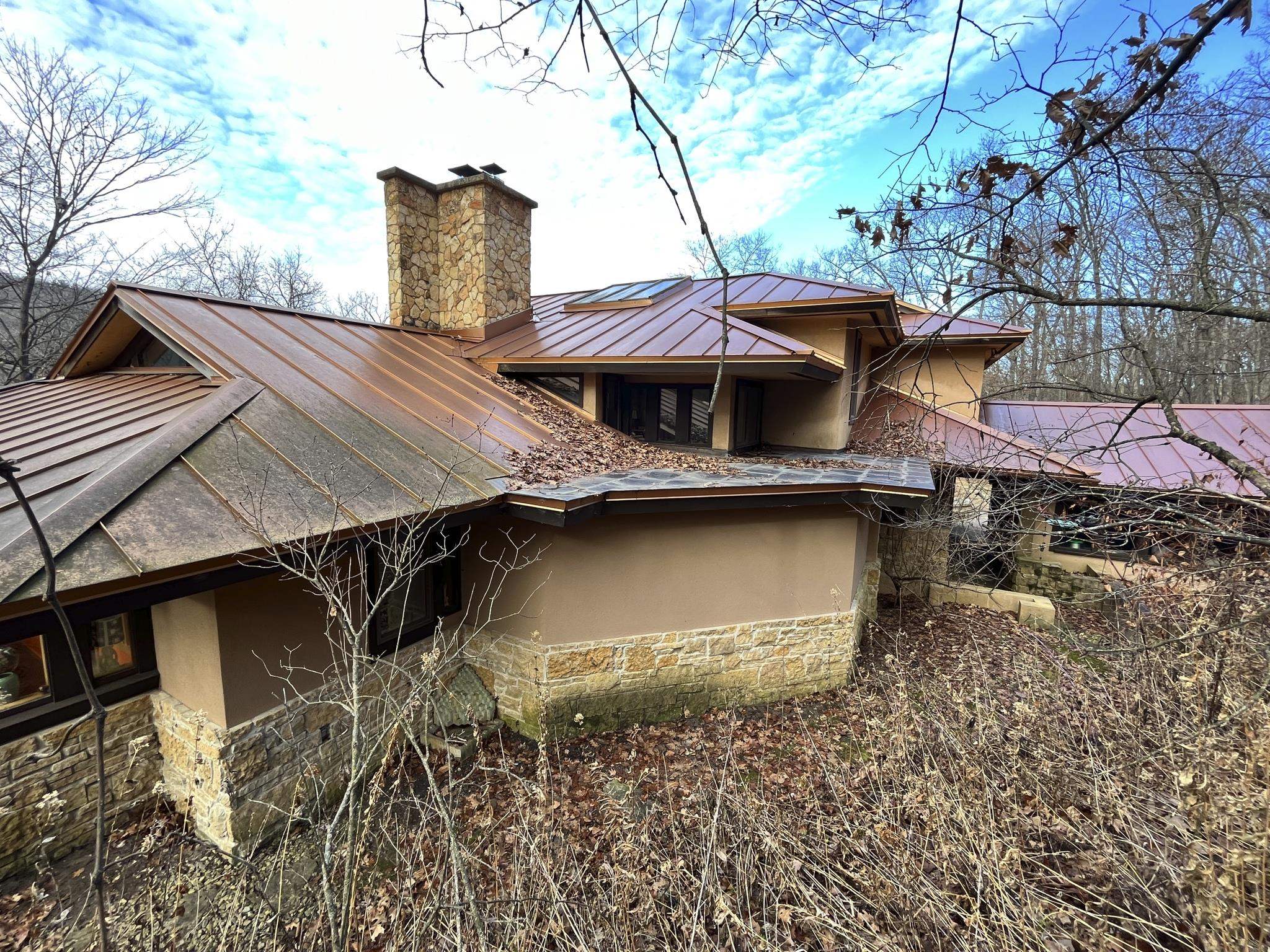 Tan wooded home with a tall stone chimney and reddish metal roof.