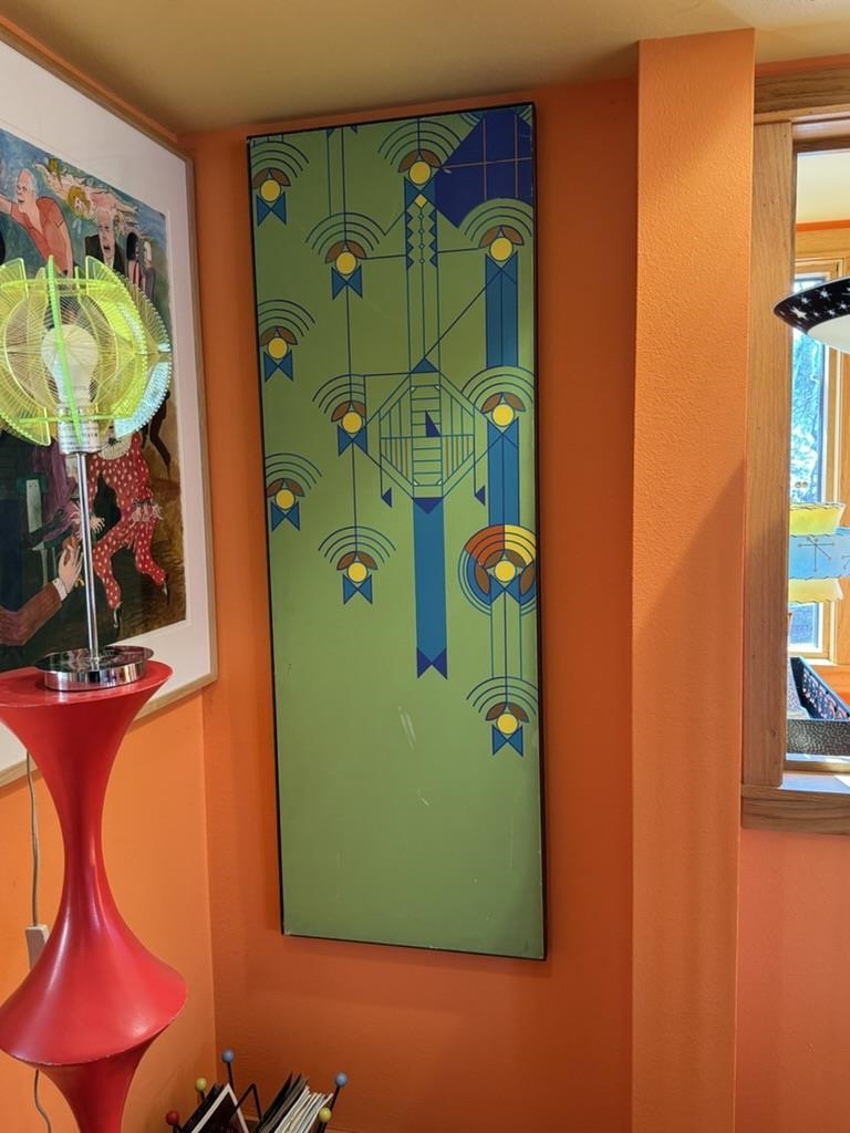 Orange interior corner with a tall geometric artwork, framed painting, and red pedestal table with a fan.