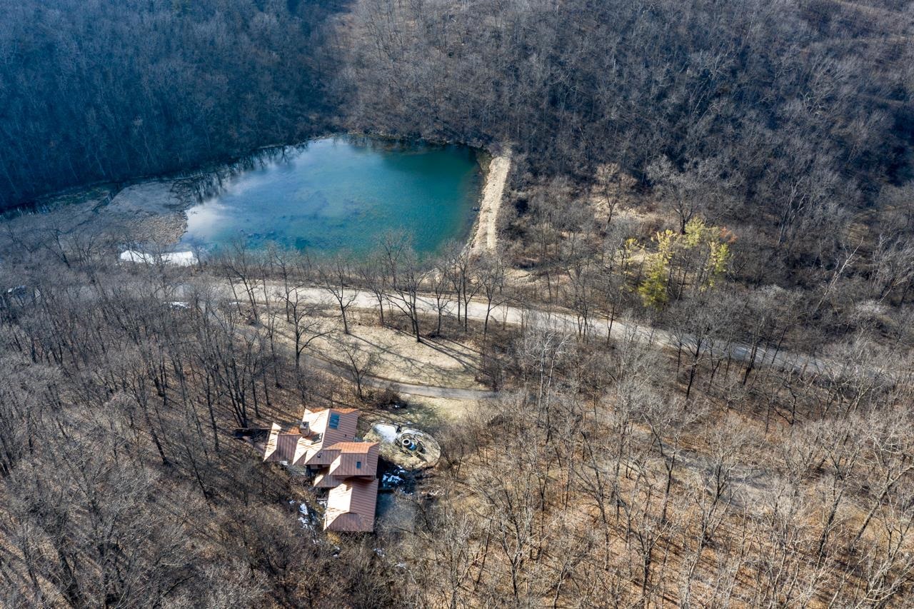 Aerial view of a wooded property with a turquoise pond and reddish-brown-roofed building.