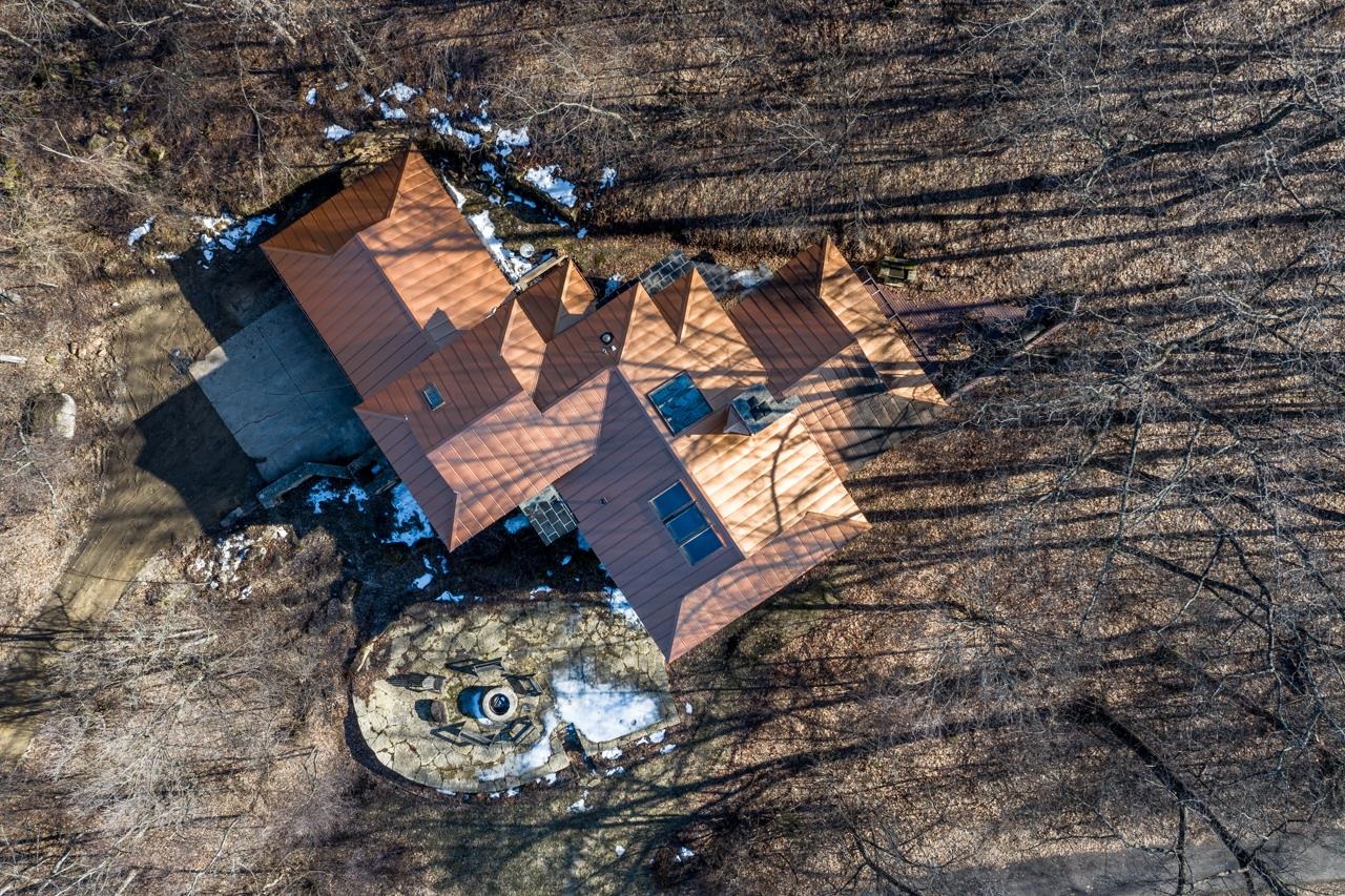 Aerial view of a reddish-brown-roofed house amid leafless woods, with a driveway and stone patio.