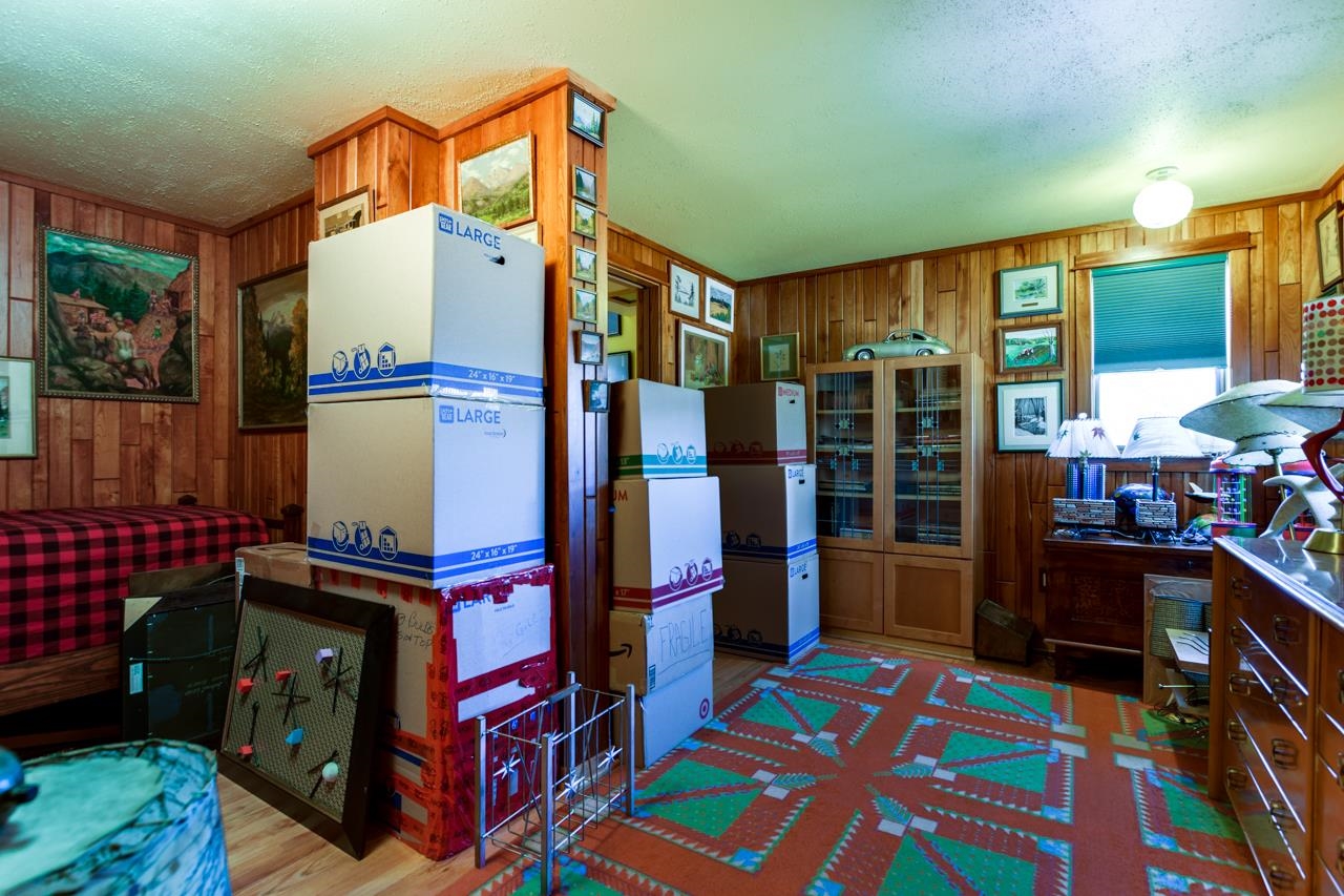Wood-paneled bedroom with a plaid bed, colorful rug, glass cabinets, and stacks of moving boxes.