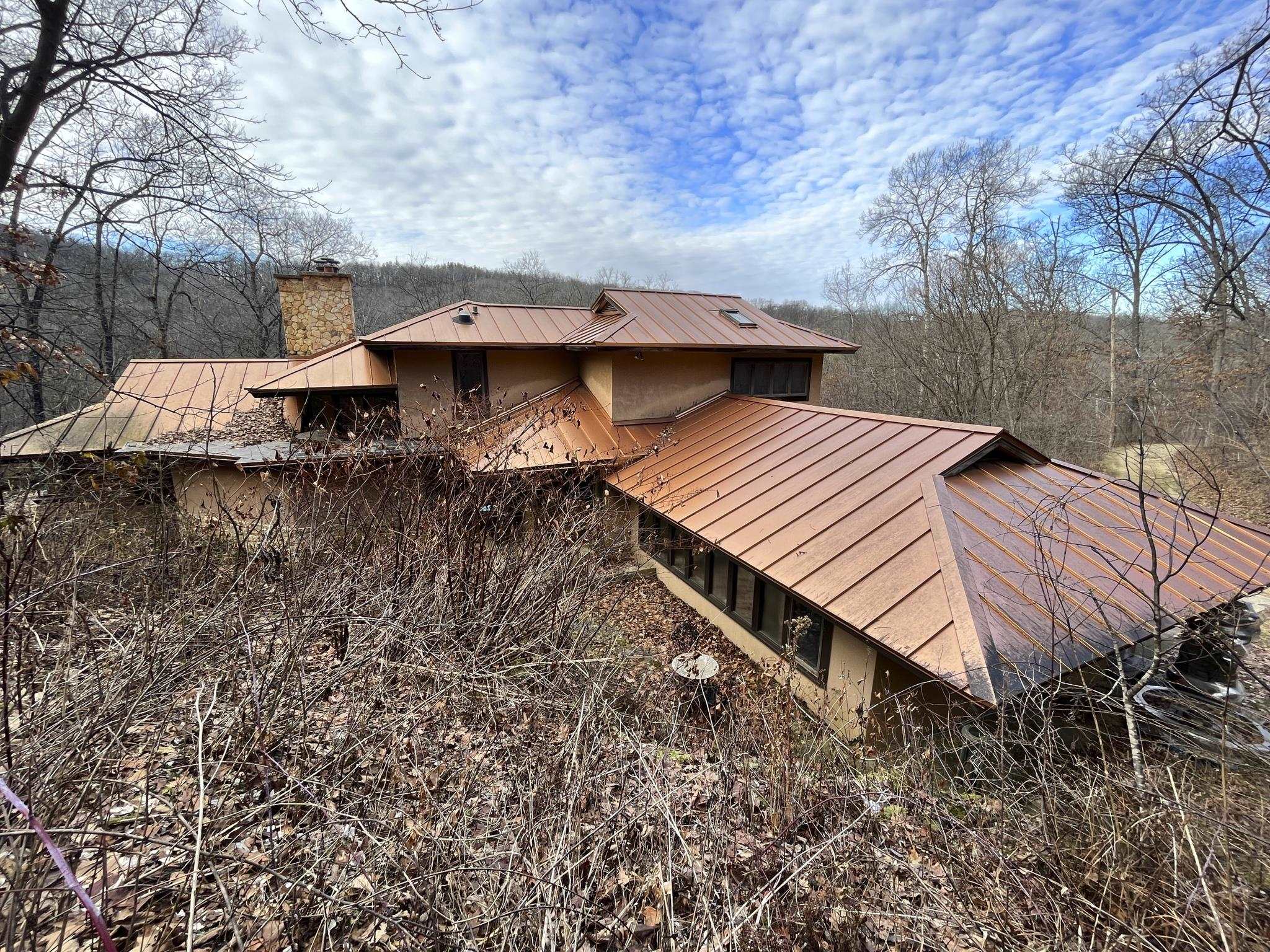 Secluded home with copper-colored metal roofs and a stone chimney among leafless trees.