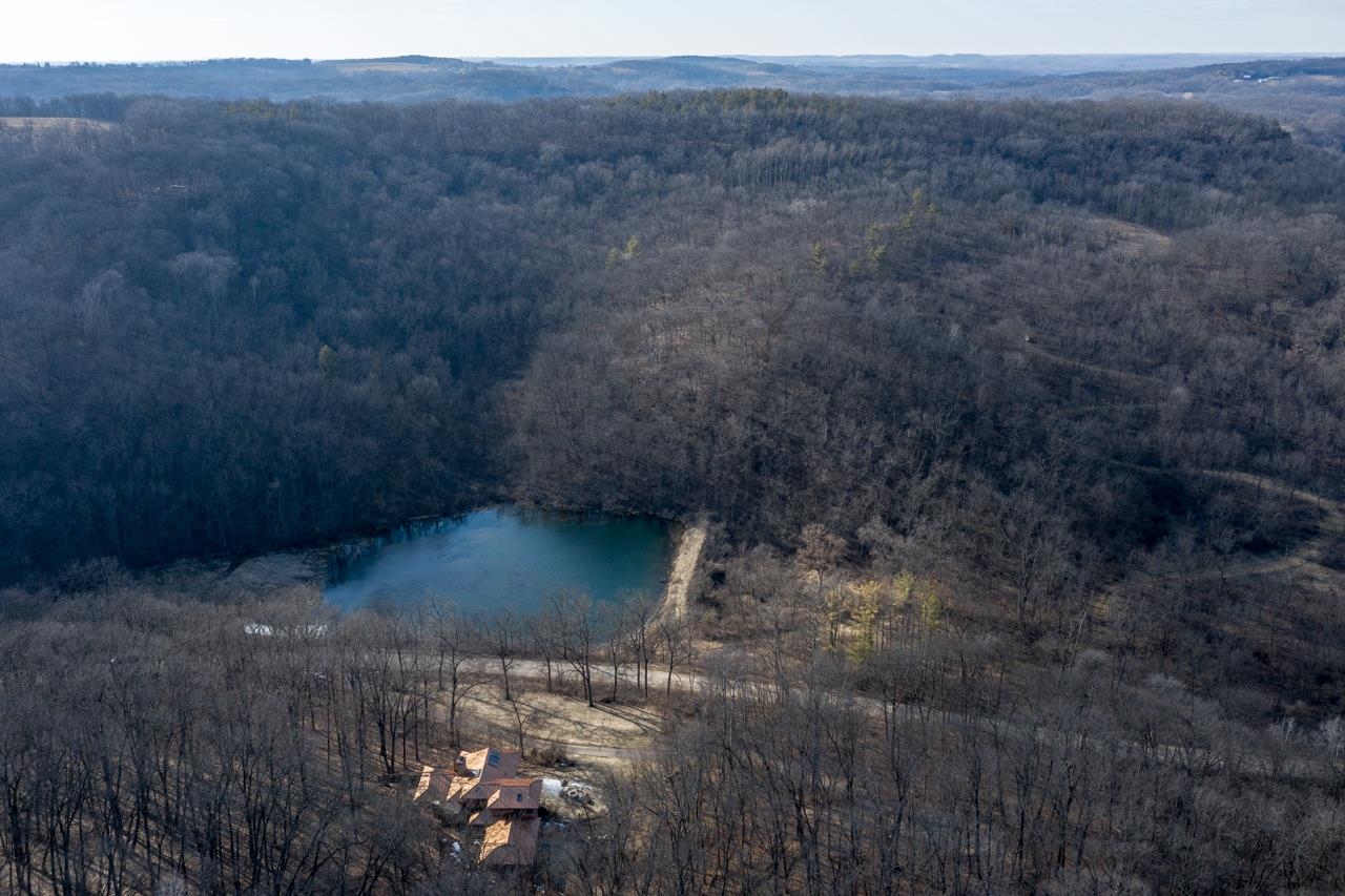 Aerial view of a wooded property with a pond, a small house, and rolling hills.