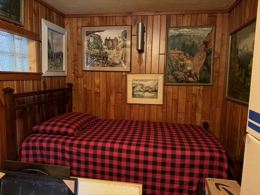 Bedroom with red-and-black plaid bedding, wood-paneled walls, and framed landscape paintings.