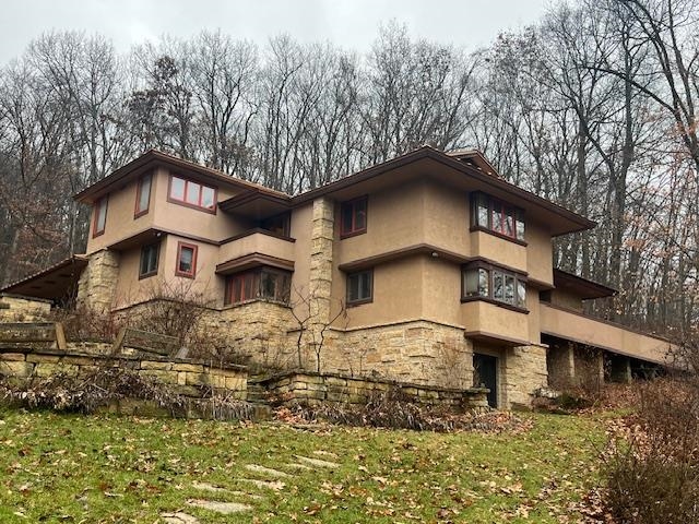 Multi-level tan home with a stone base and terraced retaining wall among leafless trees.