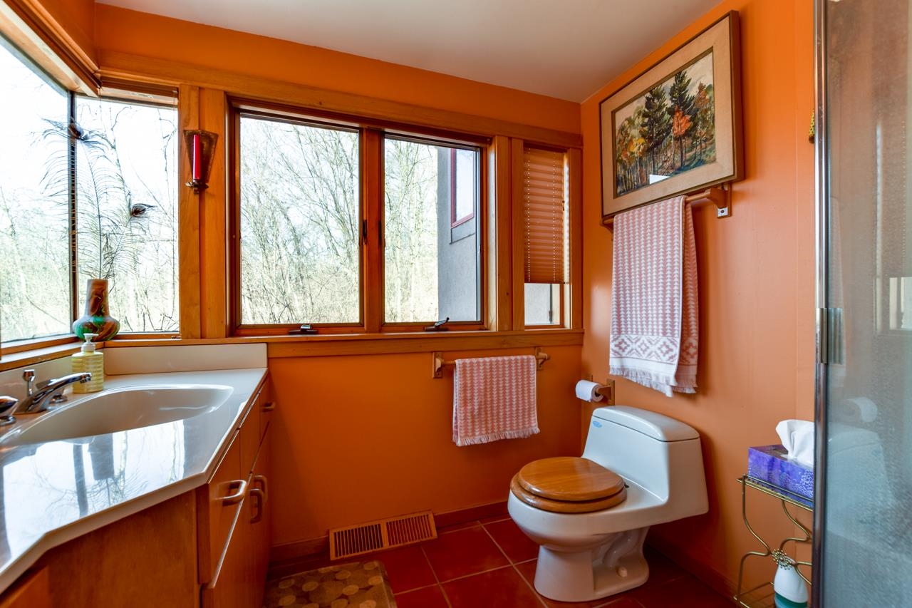 Bathroom with orange walls, a white vanity and toilet, and large windows overlooking leafless trees.