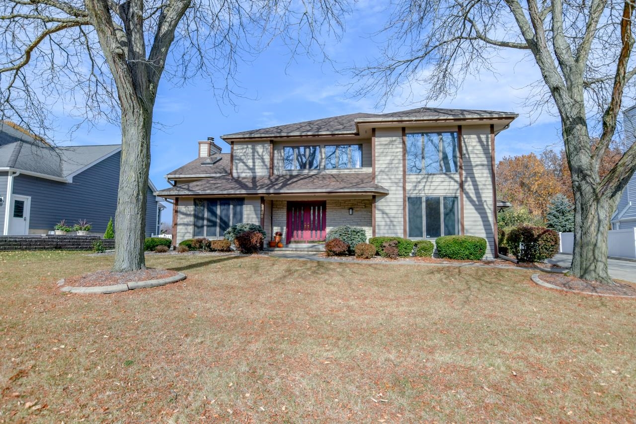 Two-story house with beige siding, stone accents, and a red front door, framed by large trees.