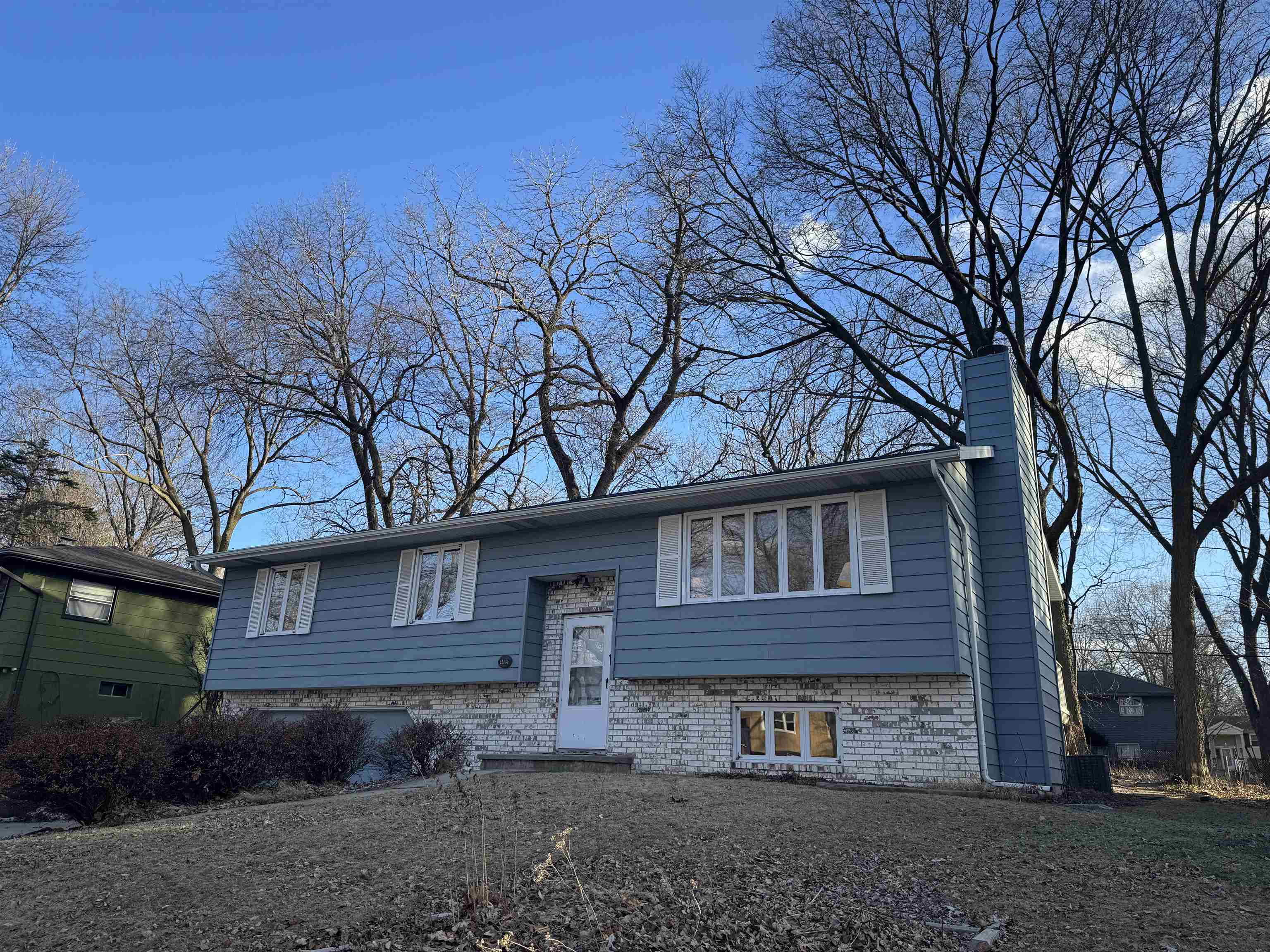 Blue split-level house with a white brick lower level and tall chimney beneath leafless trees.