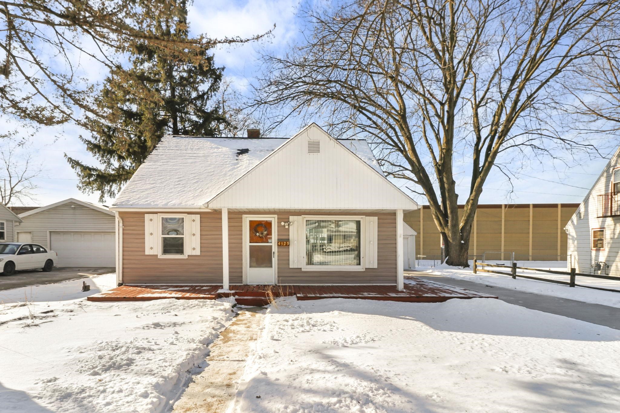 Single-story tan house with a red front deck, surrounded by snow and large bare trees.
