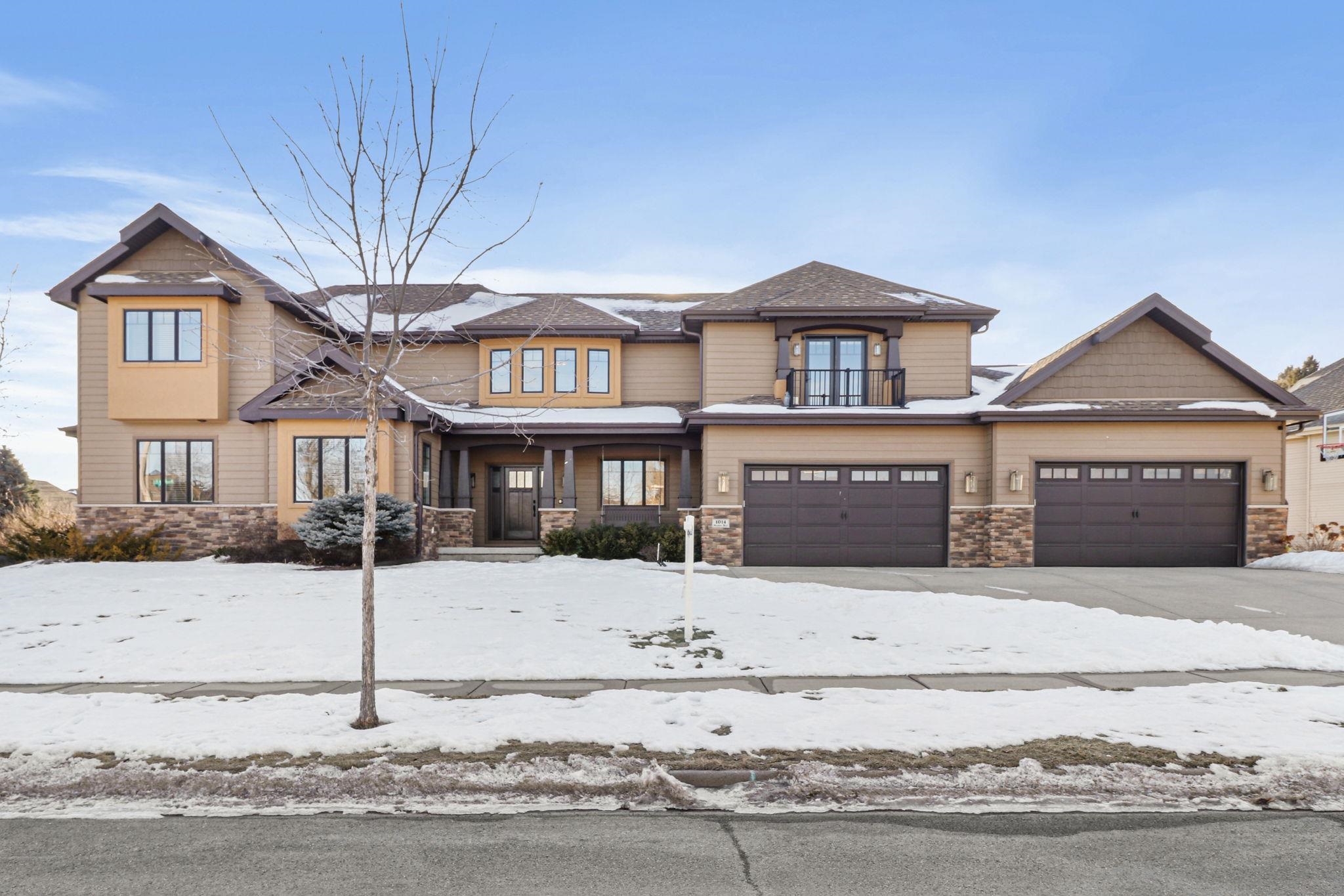 Two-story tan home with stone accents and dual garages, front yard covered in snow.