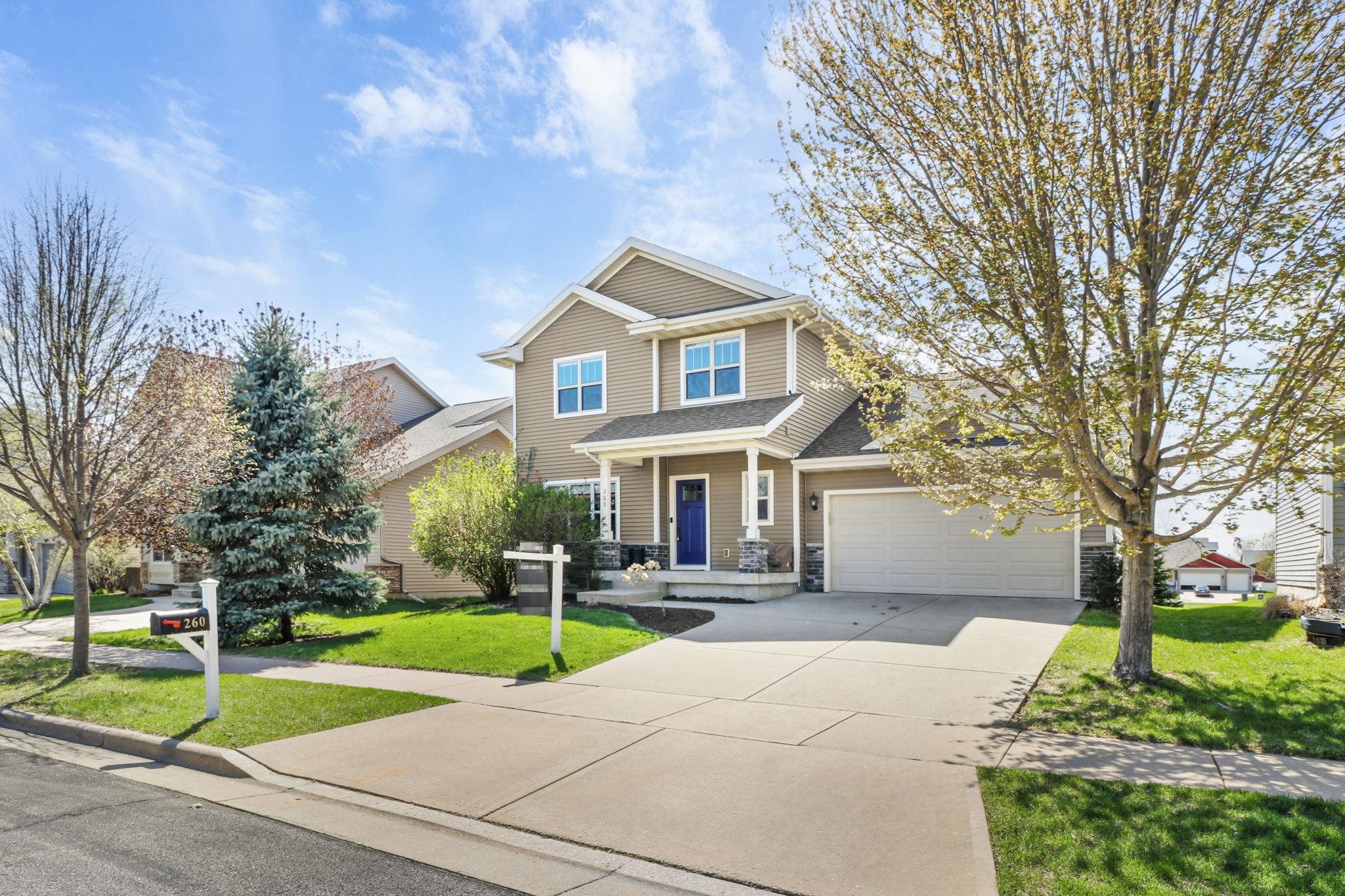 Two-story tan house with blue front door, double garage, and landscaped lawn.