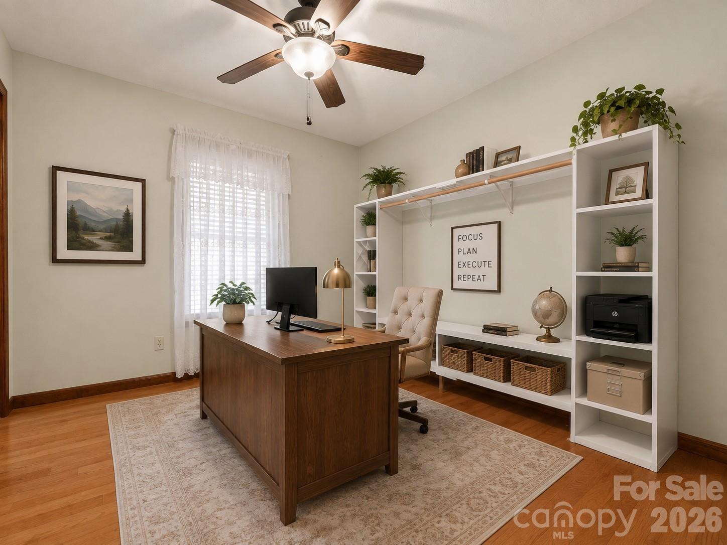 Furnished home office with a dark wood desk, white built-in shelving, and a curtained window.