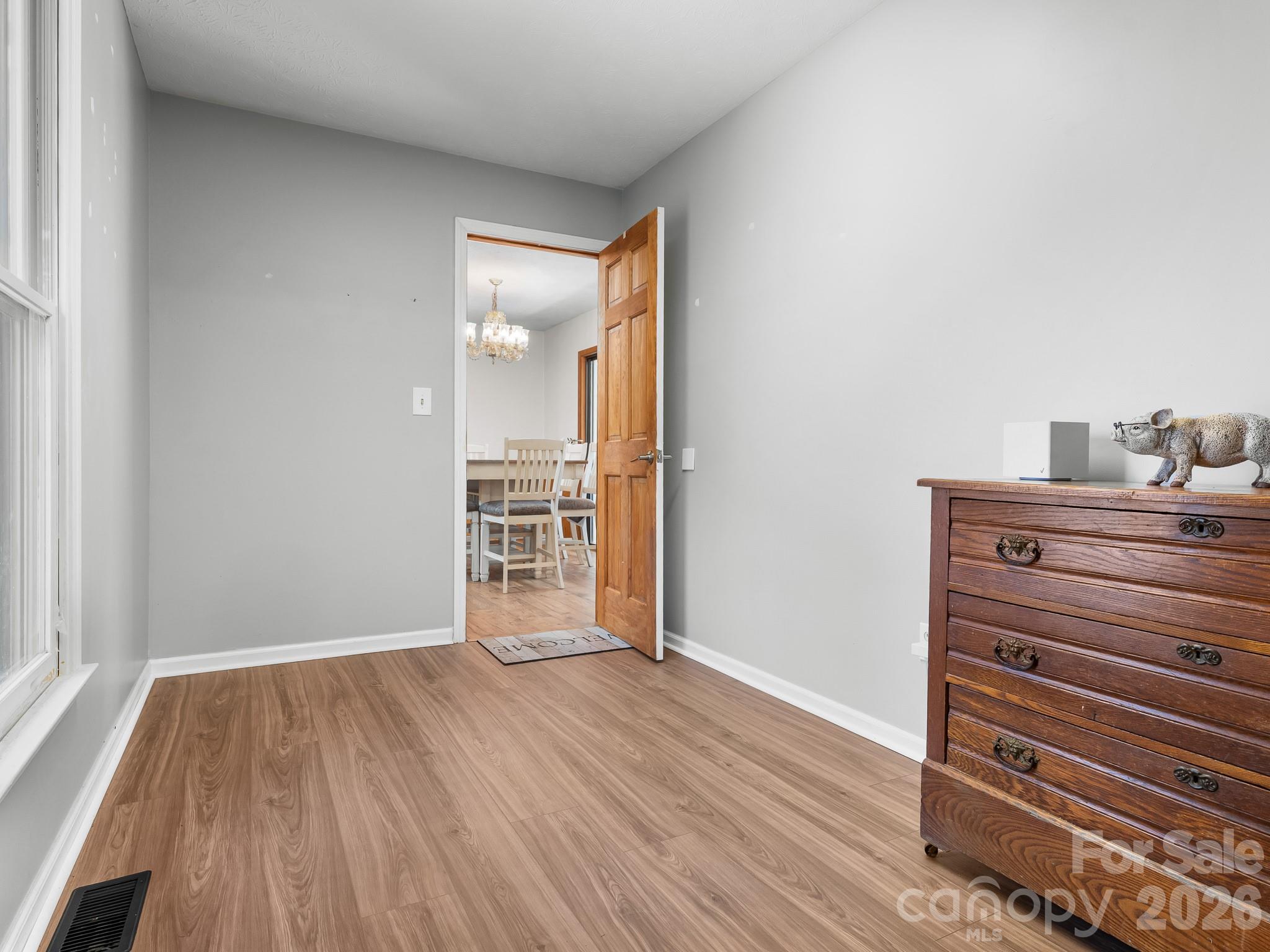 Bright gray room with wood-look floors, a tall window, and a dark wood dresser beside an open door.