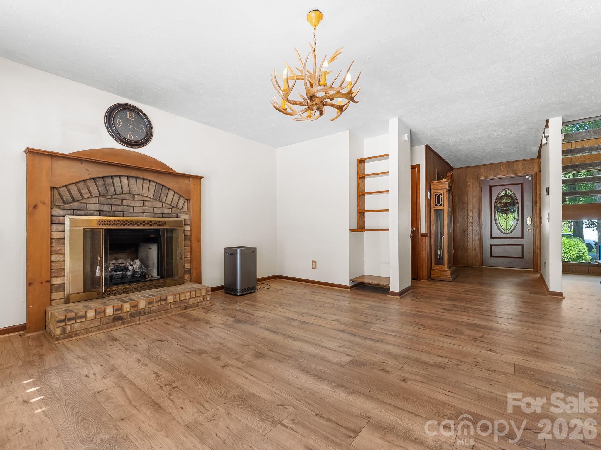 Empty living room with a brick fireplace, wood-look floors, and antler chandelier.