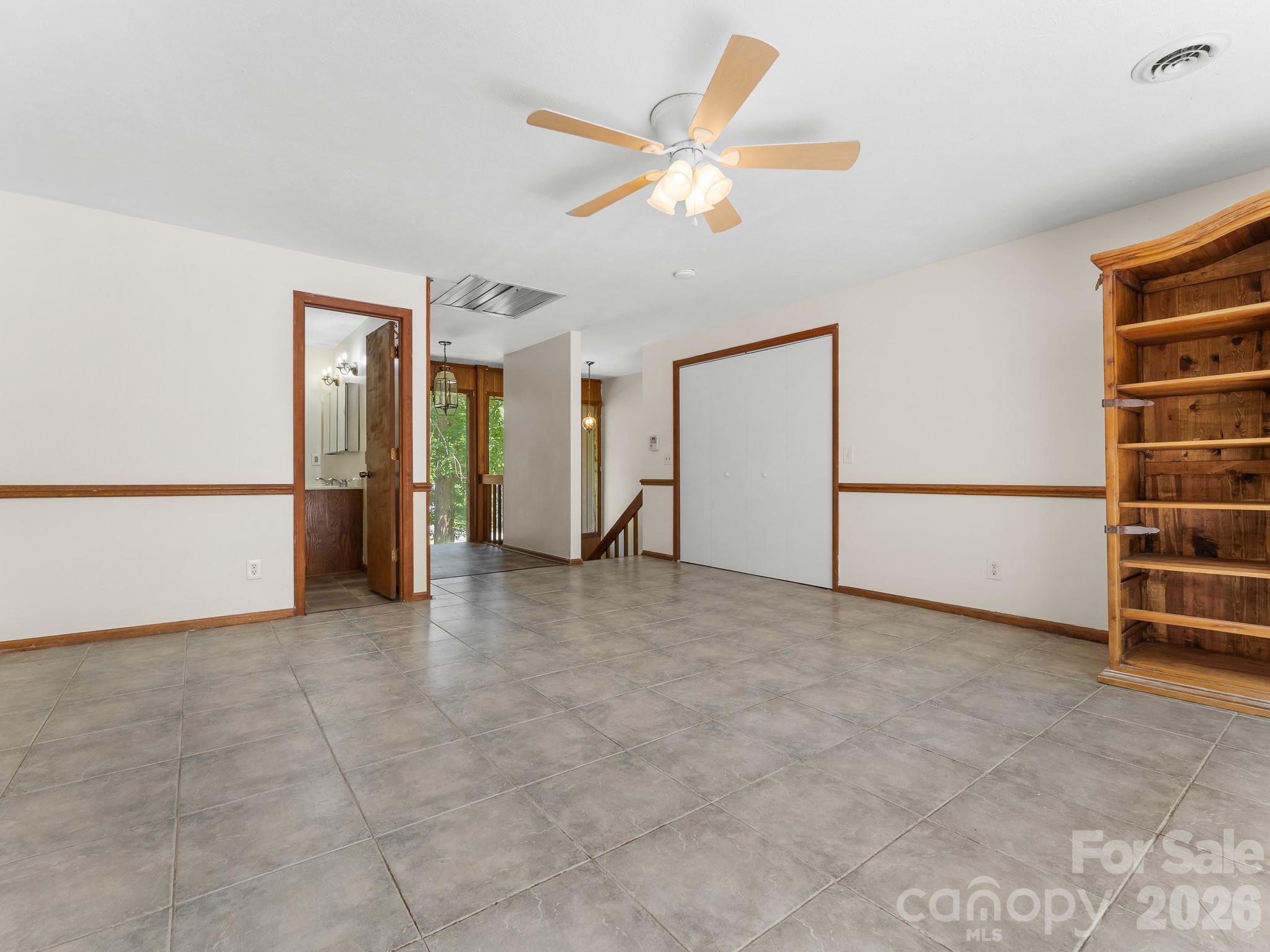 Empty tiled room with a ceiling fan, built-in wooden bookcase, and doorway to a bathroom.
