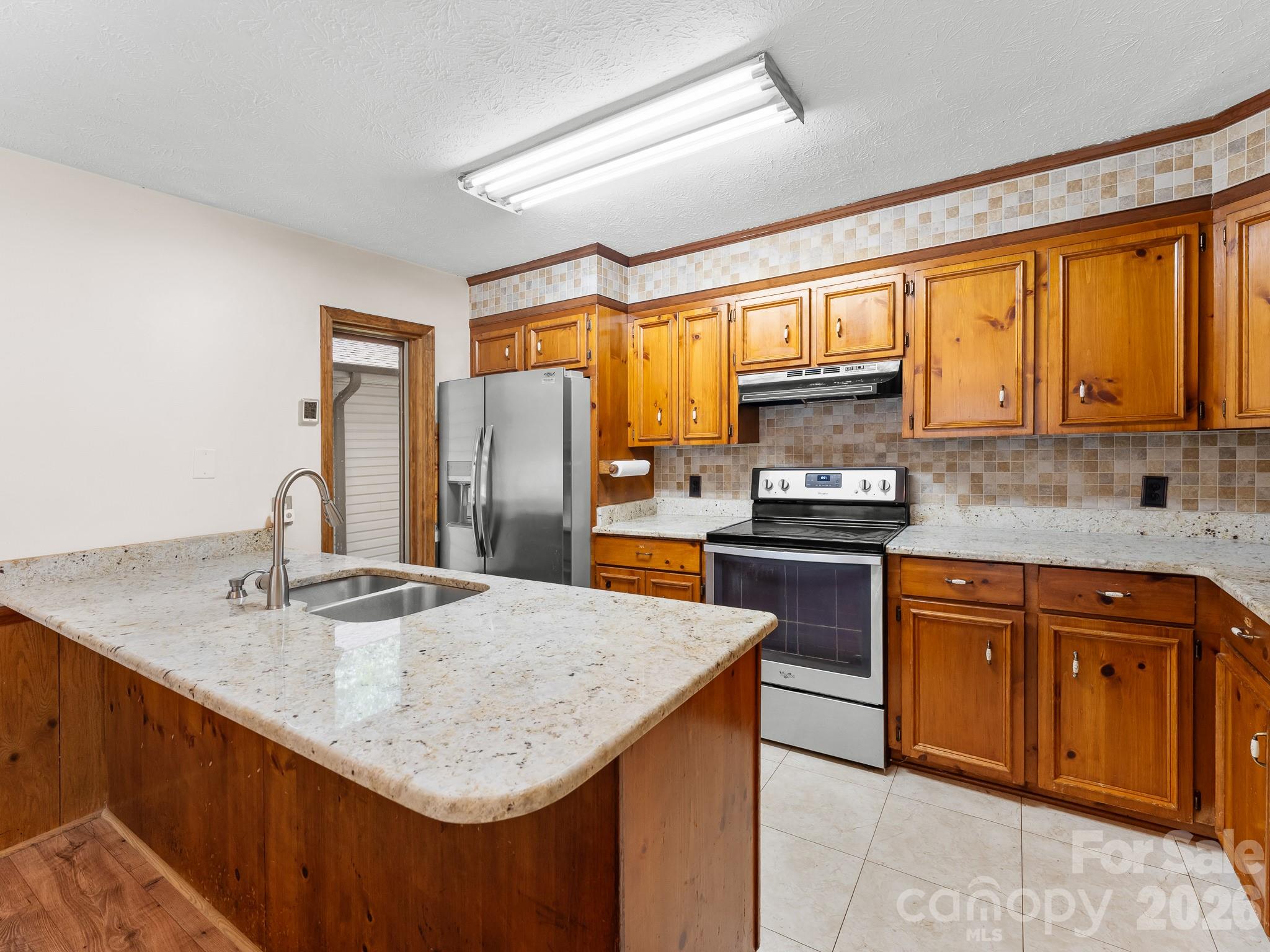 Kitchen with honey-toned wood cabinets, stone counters, a large sink peninsula, and stainless steel appliances.