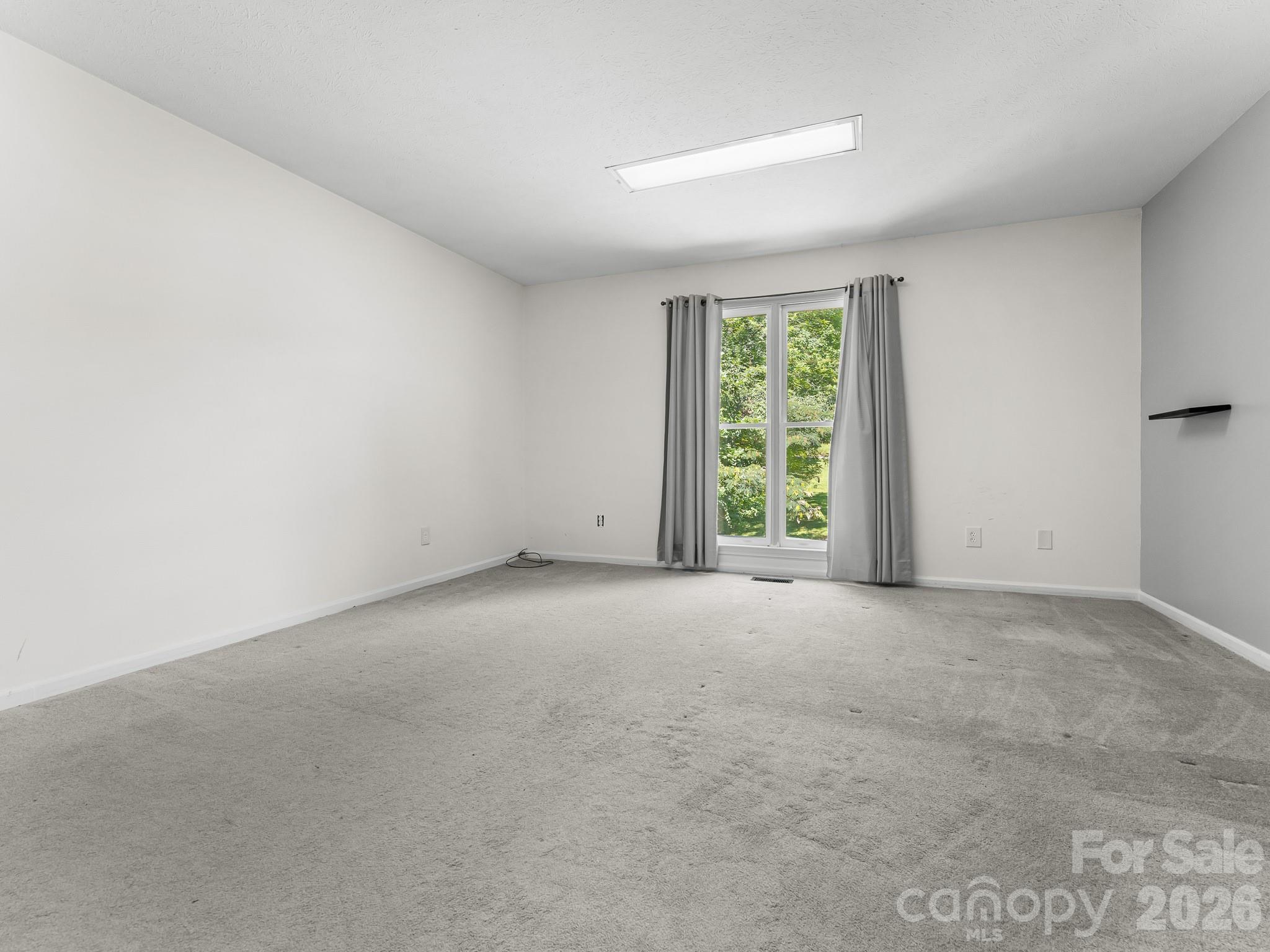 Empty carpeted room with gray curtains framing a large window overlooking leafy greenery.