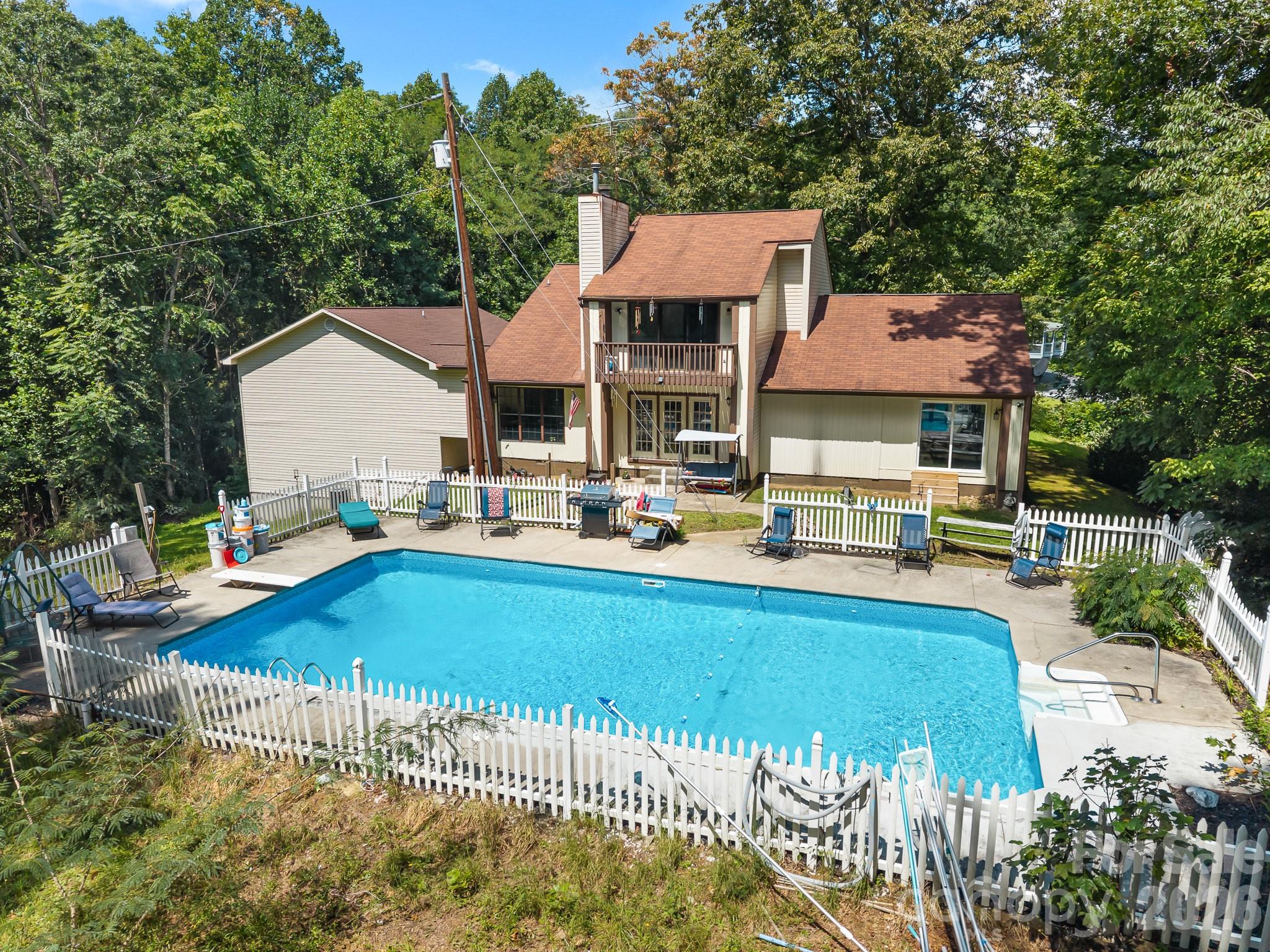 Two-story house behind a large fenced swimming pool, with a wooded backdrop and patio seating.