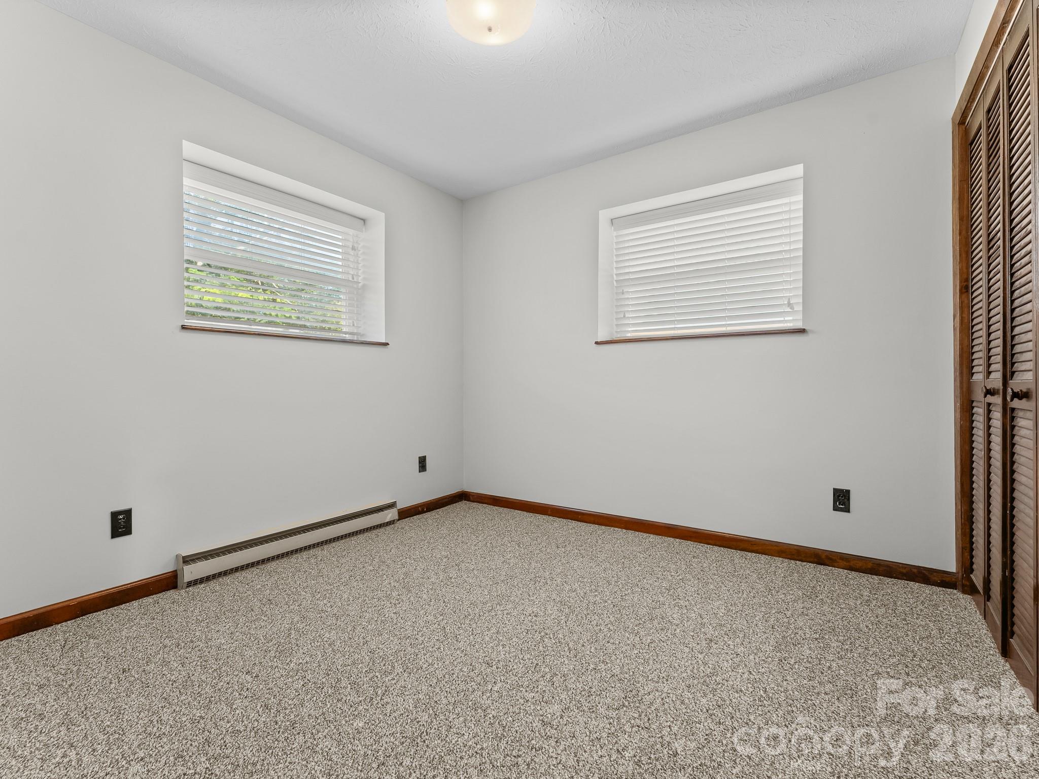 Empty bedroom with gray carpet, two windows, a baseboard heater, and wooden louvered closet doors.