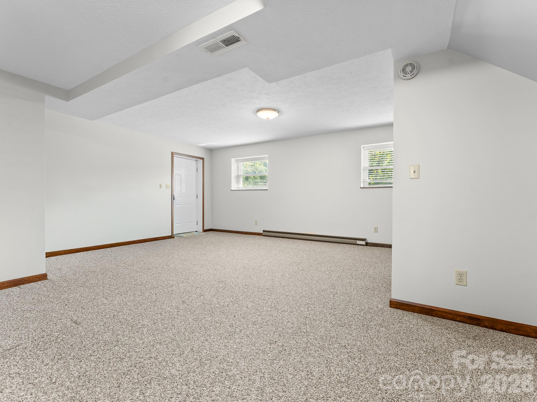 Empty carpeted room with two windows, a white door, and a baseboard heater.