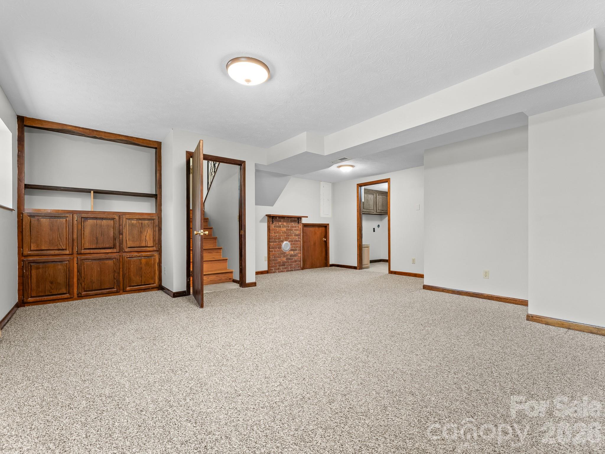 Unfurnished carpeted room with built-in wood cabinets, a staircase, and a small brick fireplace.