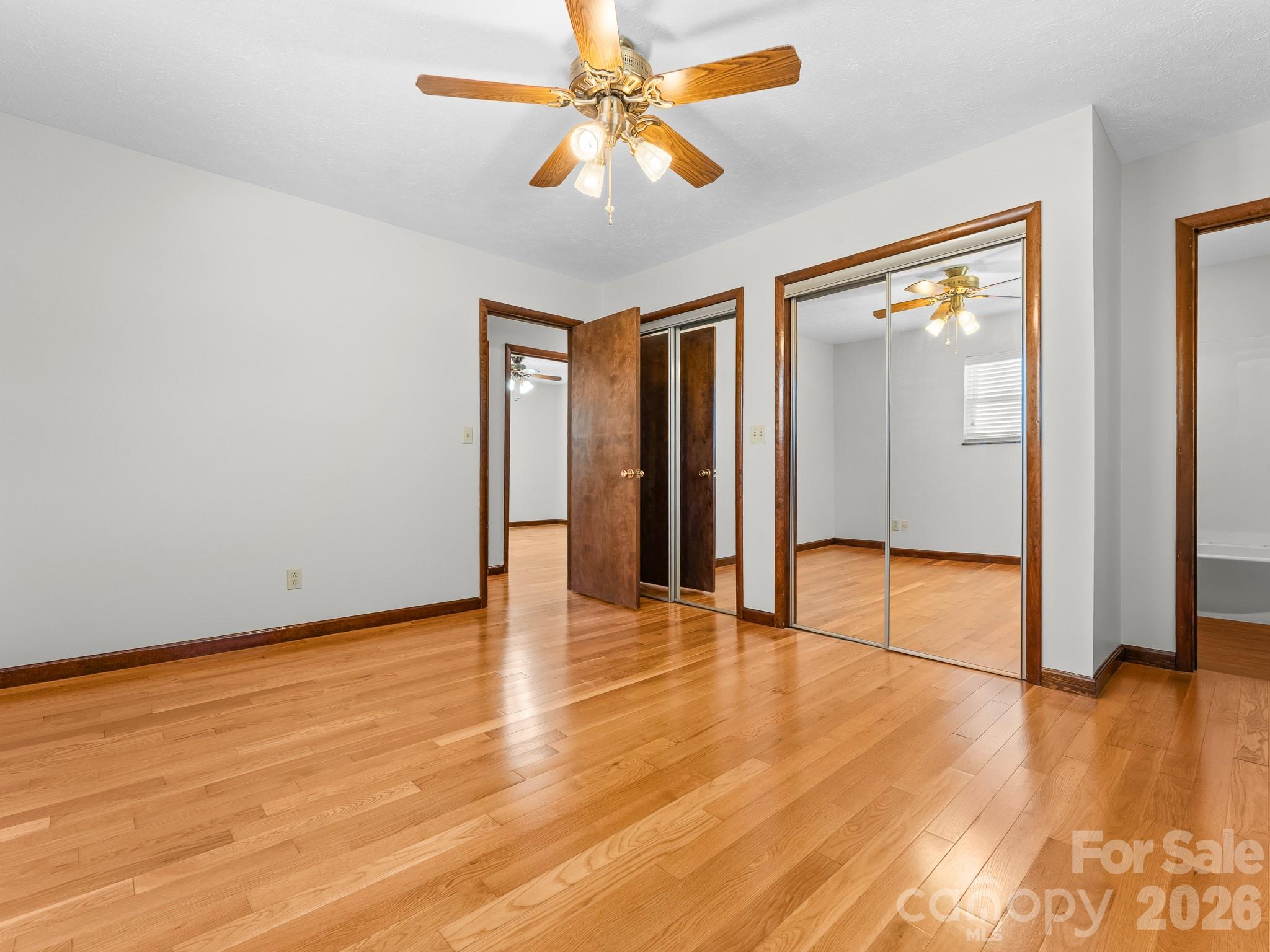 Empty bedroom with hardwood floors, mirrored closet doors, and a wood-blade ceiling fan.