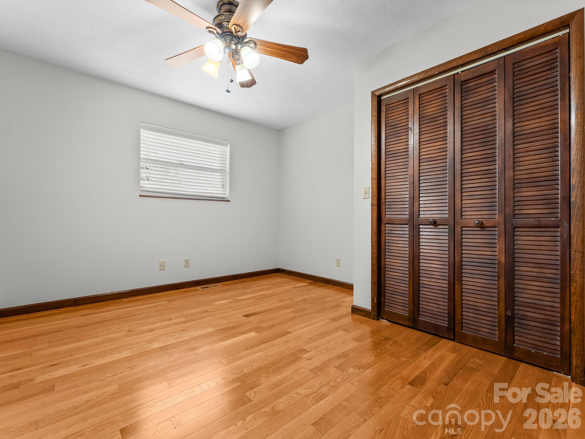 Empty bedroom with hardwood floors, a ceiling fan, and dark louvered closet doors.