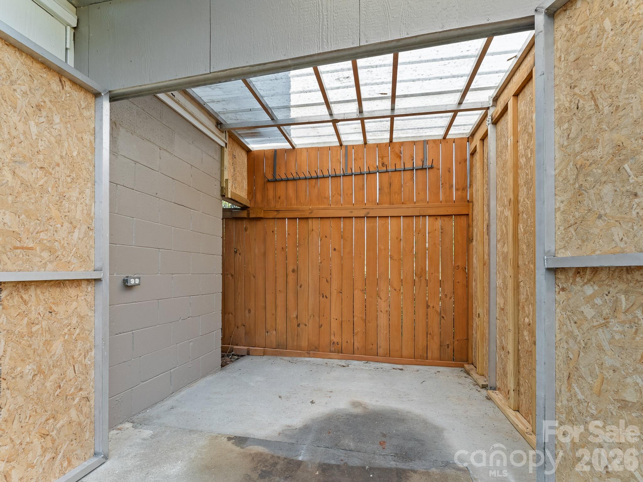 Covered parking area with a translucent roof, concrete floor, and wooden rear wall.