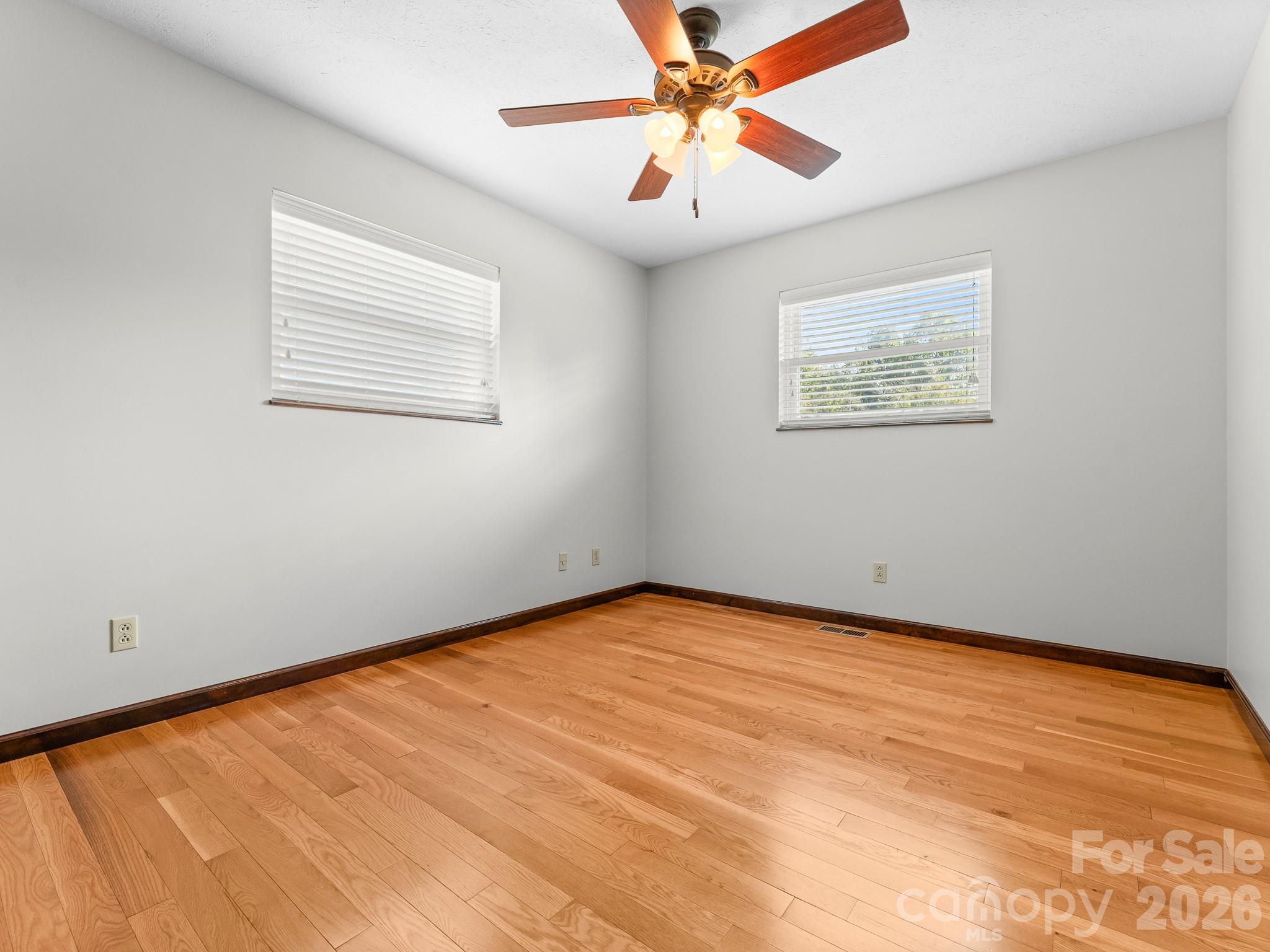 Empty room with light hardwood floors, two windows with blinds, and a lit ceiling fan.