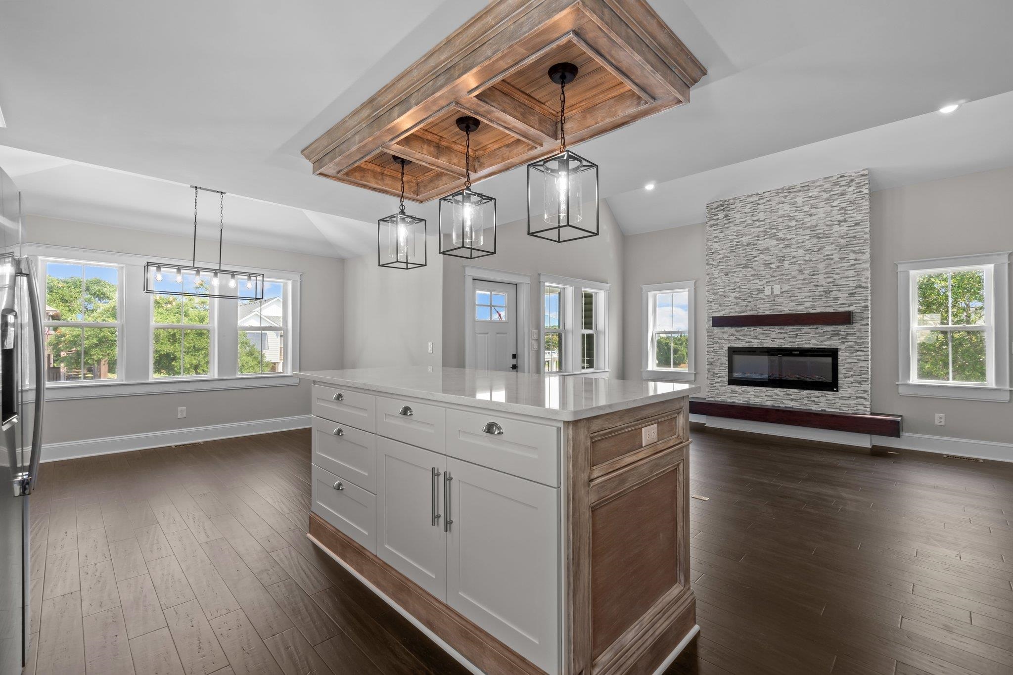 Open-plan kitchen and living area with a white island, wood ceiling feature, and stone fireplace.