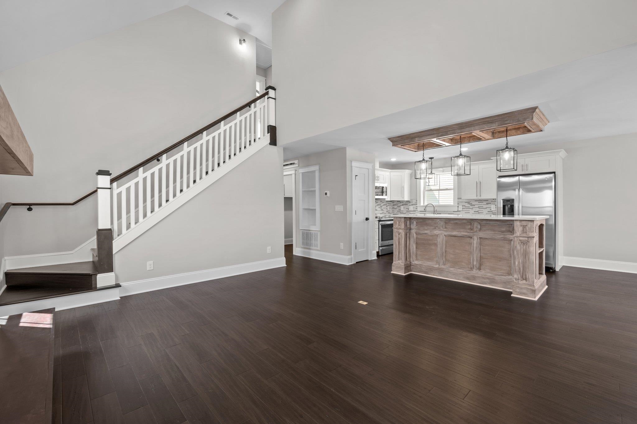 Open living area with a large kitchen island, stainless appliances, dark flooring, and staircase.