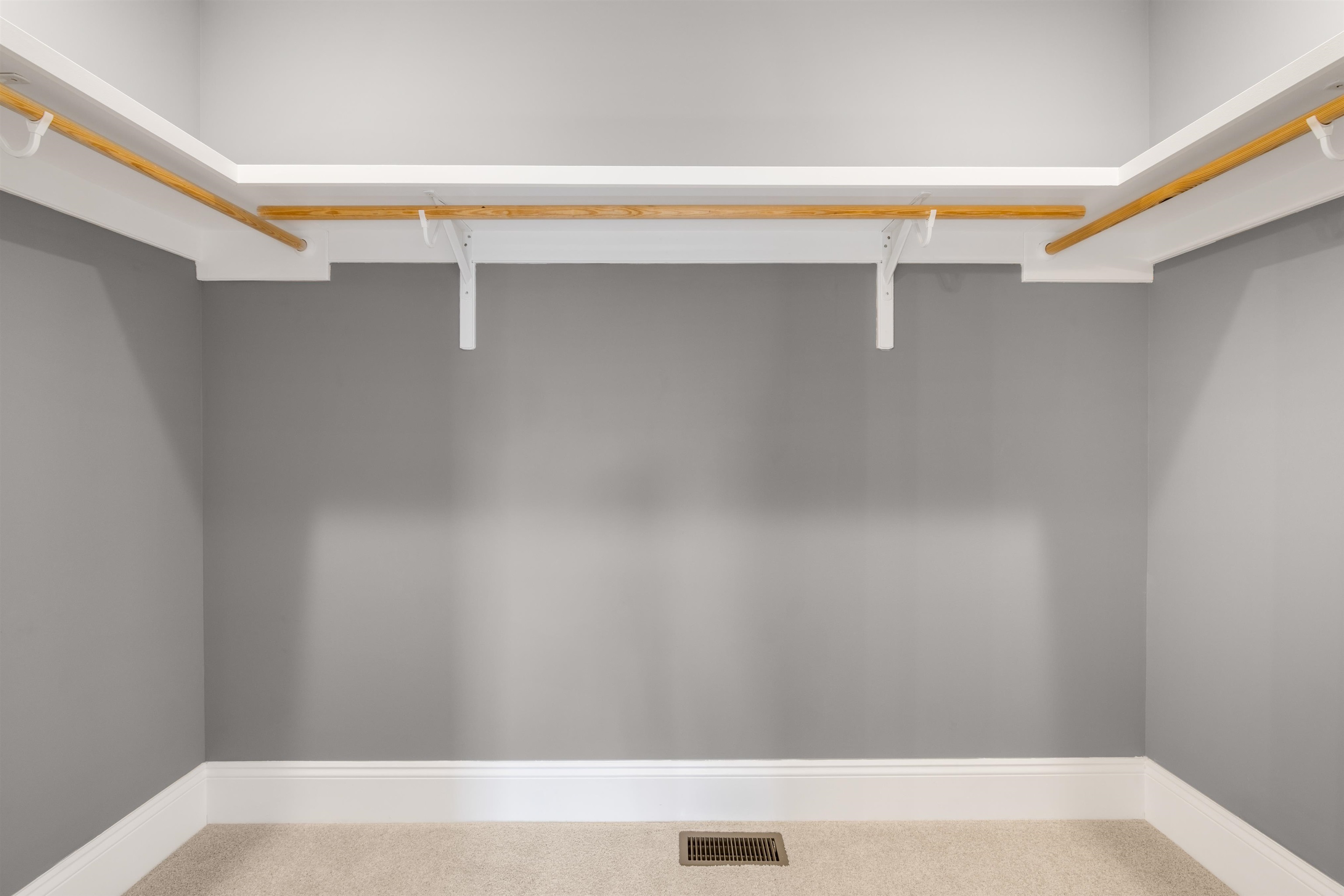 Empty walk-in closet with wooden hanging rods, gray walls, and beige carpet.