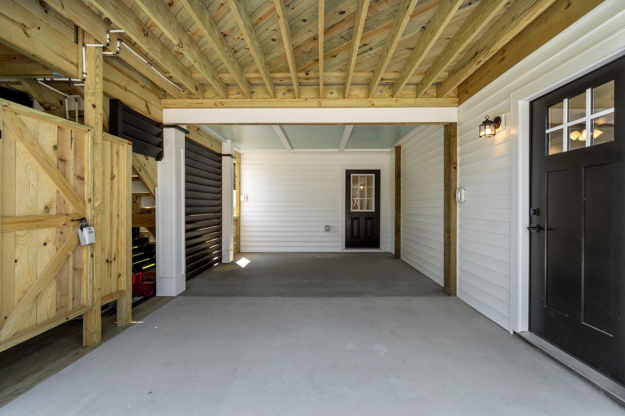 Covered parking area with exposed timber beams, a wooden staircase, and black entry doors.