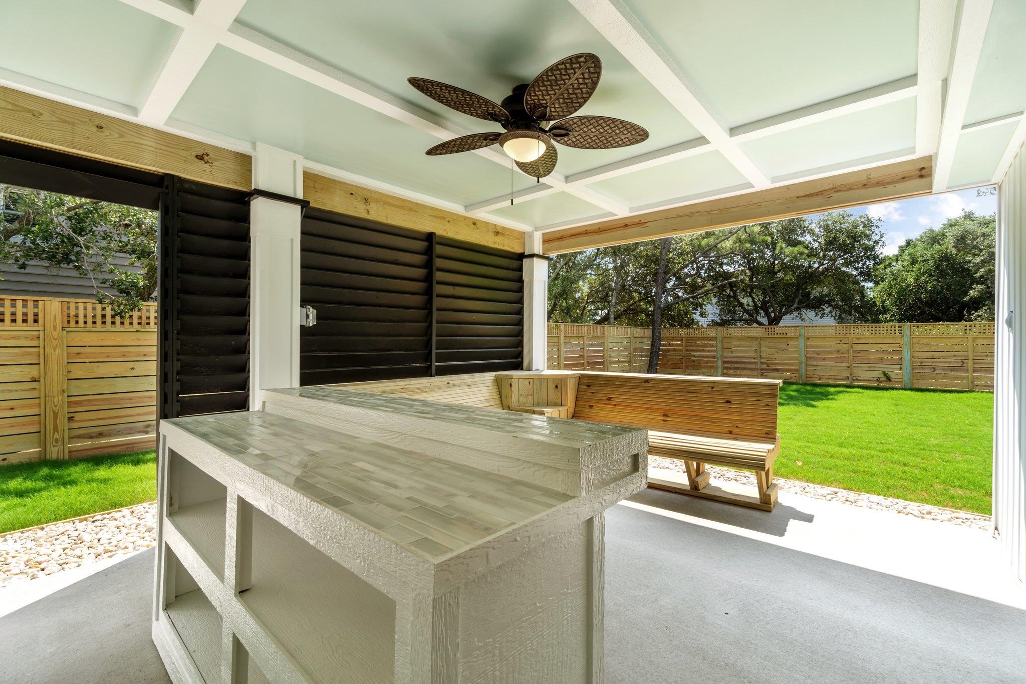 Covered patio with tiled outdoor counter, ceiling fan, and wooden bench beside a fenced lawn.