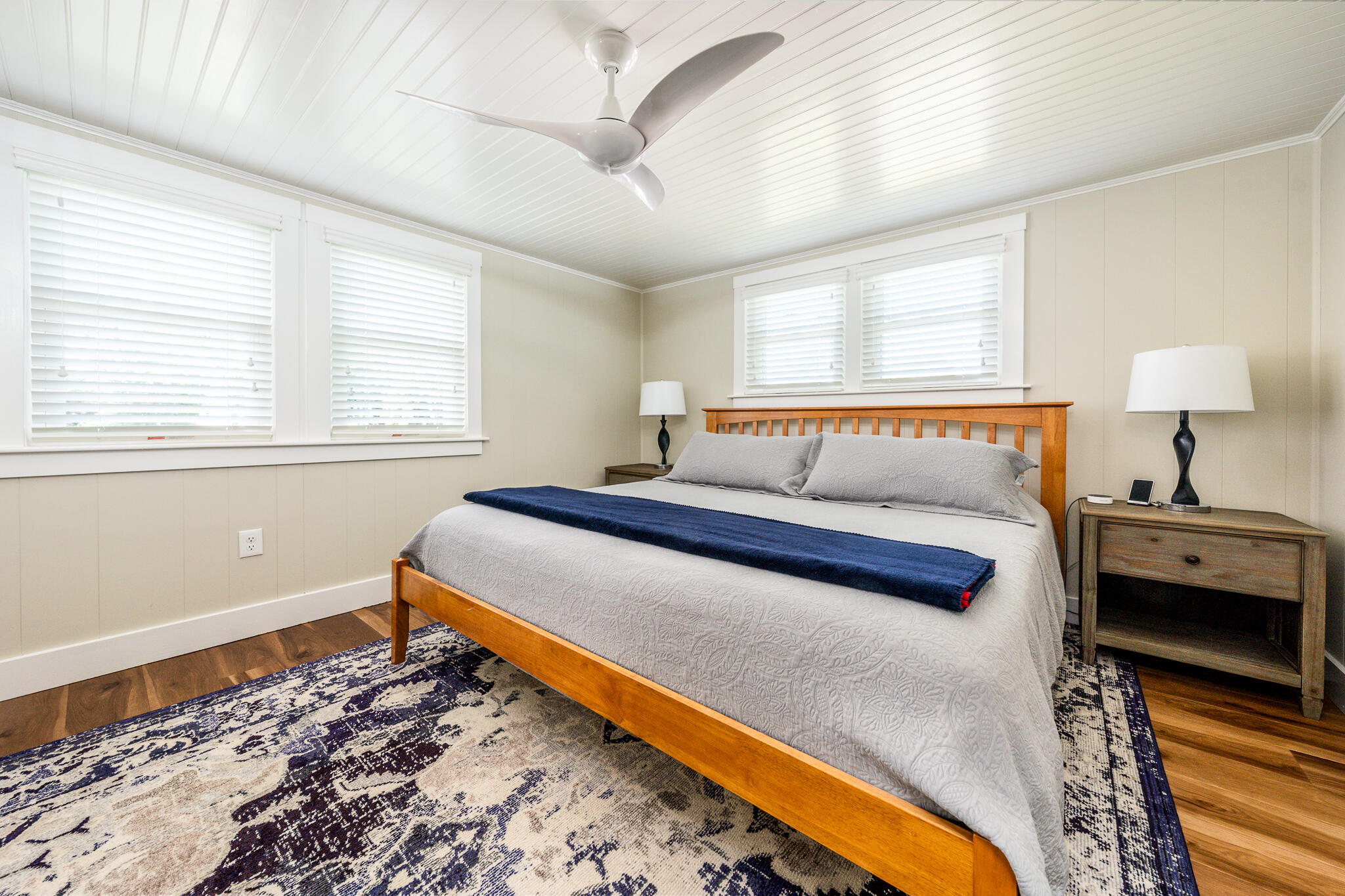 Bedroom with a wooden bed, navy throw, patterned rug, and white-blinded windows.