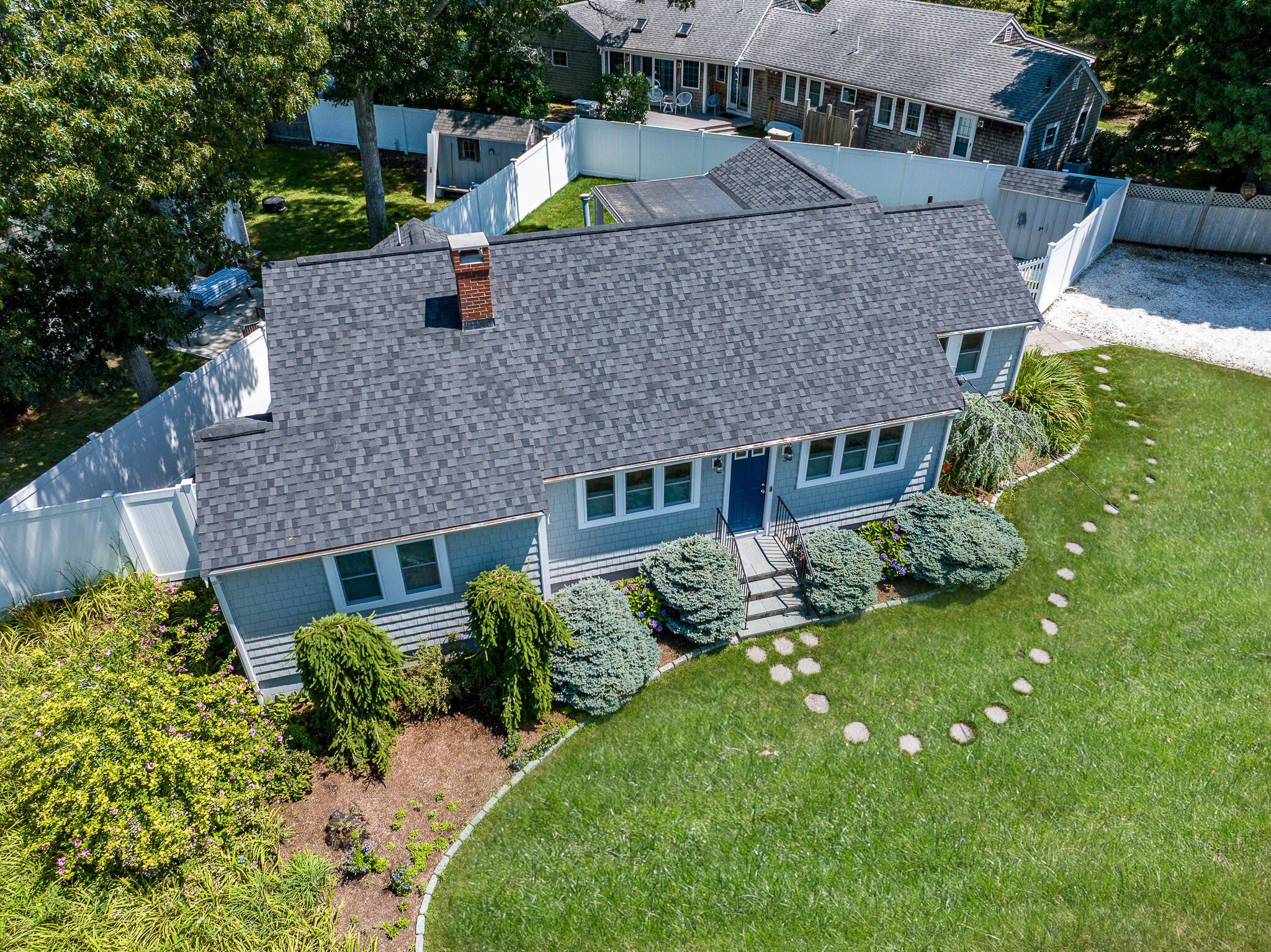 Aerial view of a blue-gray home with landscaped gardens, a white fenced yard, and lawn stepping stones.