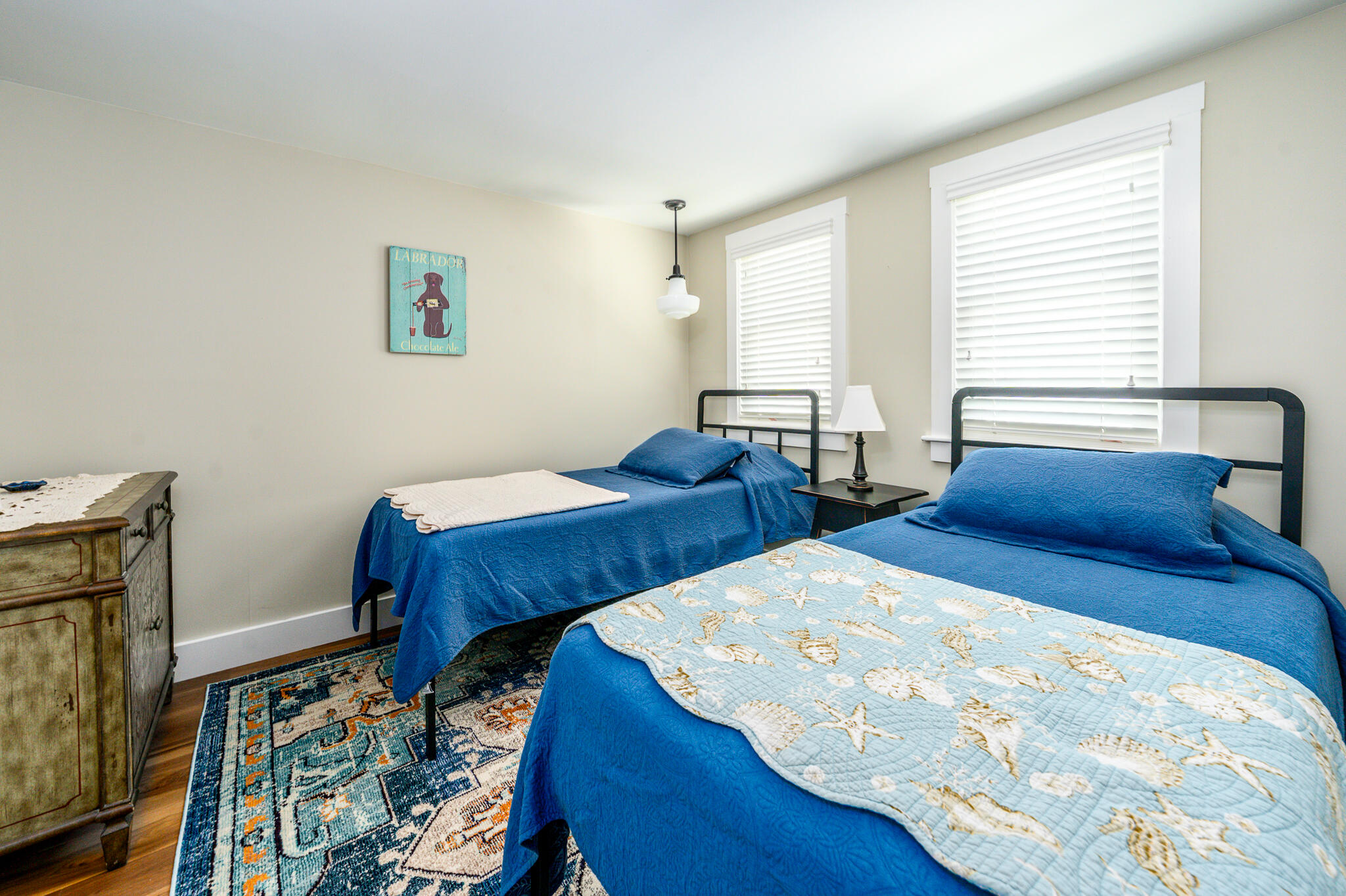 Bedroom with two blue-covered beds, white blinds, and a patterned rug.