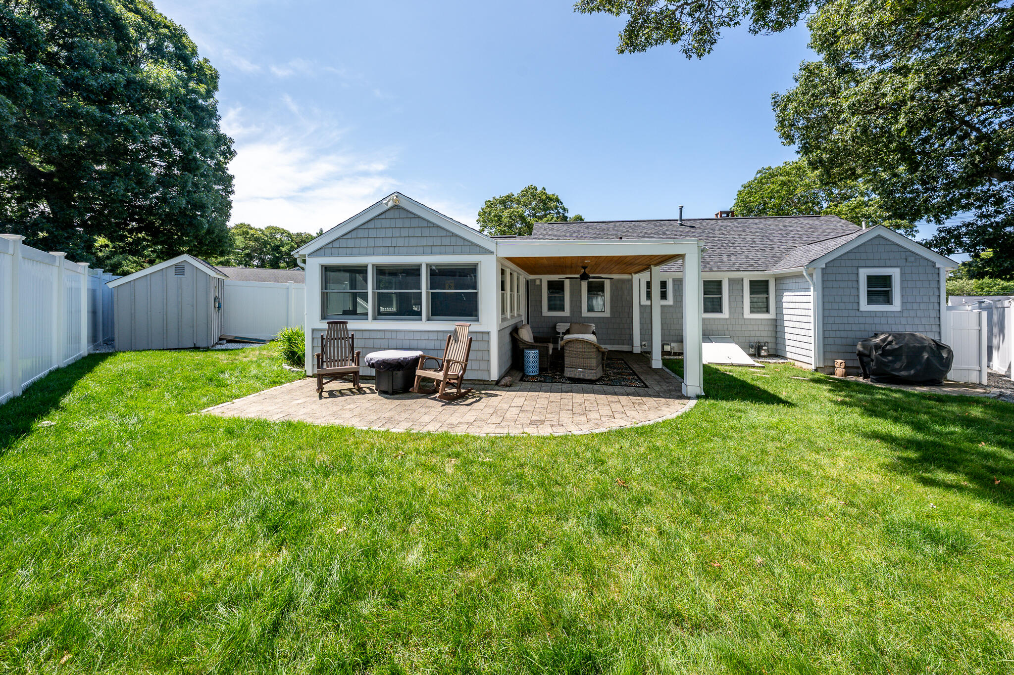 Gray shingle-sided home with a covered patio and paver seating area overlooking a fenced lawn.