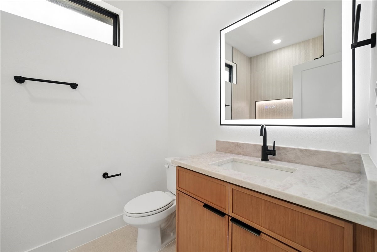 Modern bathroom with a wood-front vanity, pale stone counter, large lit mirror, and white toilet.