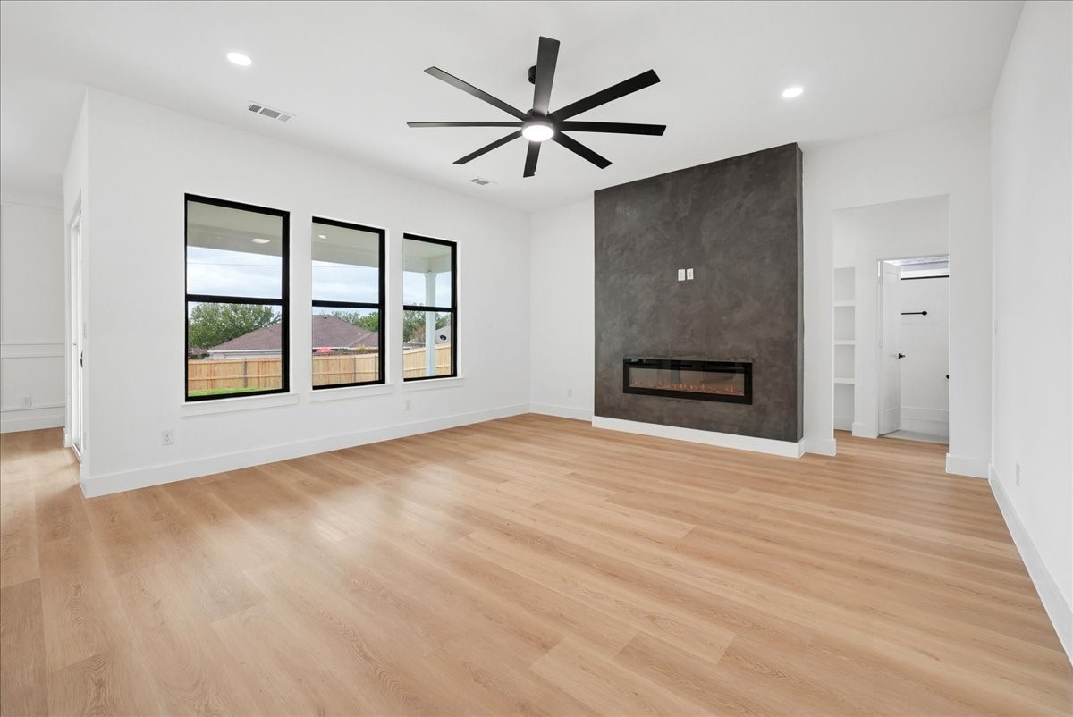 Empty living room with a dark fireplace wall, three windows, light wood floors, and a black ceiling fan.