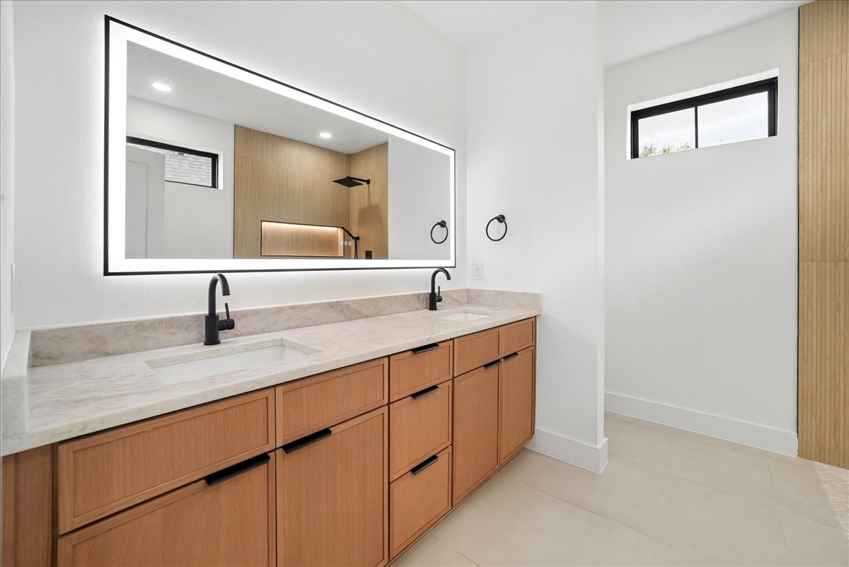 Modern bathroom with a double vanity, light wood cabinets, pale stone counters, and a large mirror.