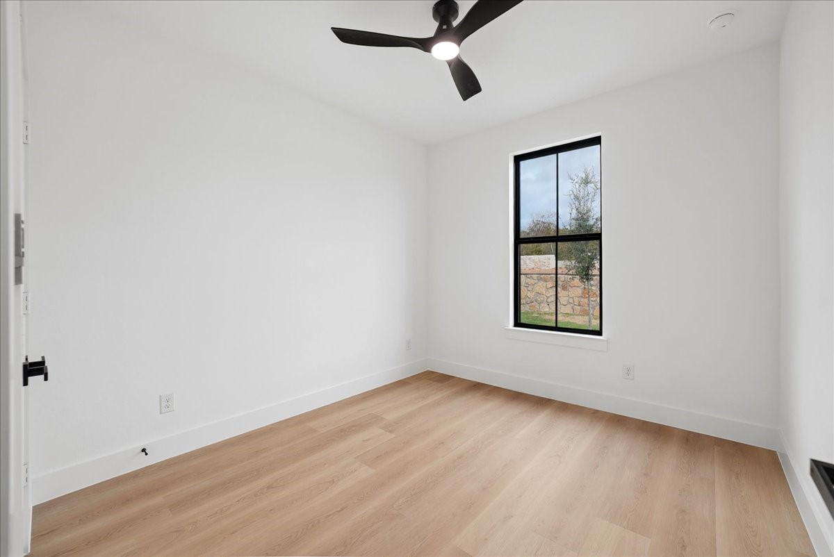 Empty room with light wood floors, a black ceiling fan, and a window overlooking a stone wall.