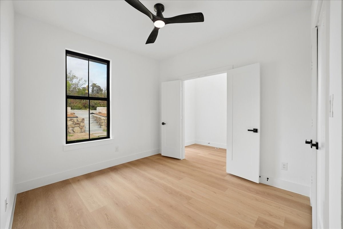 Empty bedroom with light wood floors, a black-framed window, and open double doors to an adjoining room.